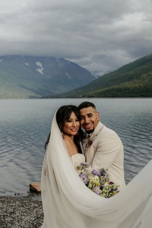 Wedding portraits by a mountain lake with flowing veil and purple bouquet.