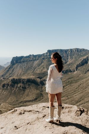 A person in a white dress and boots stands on a cliff edge overlooking desert mountains.