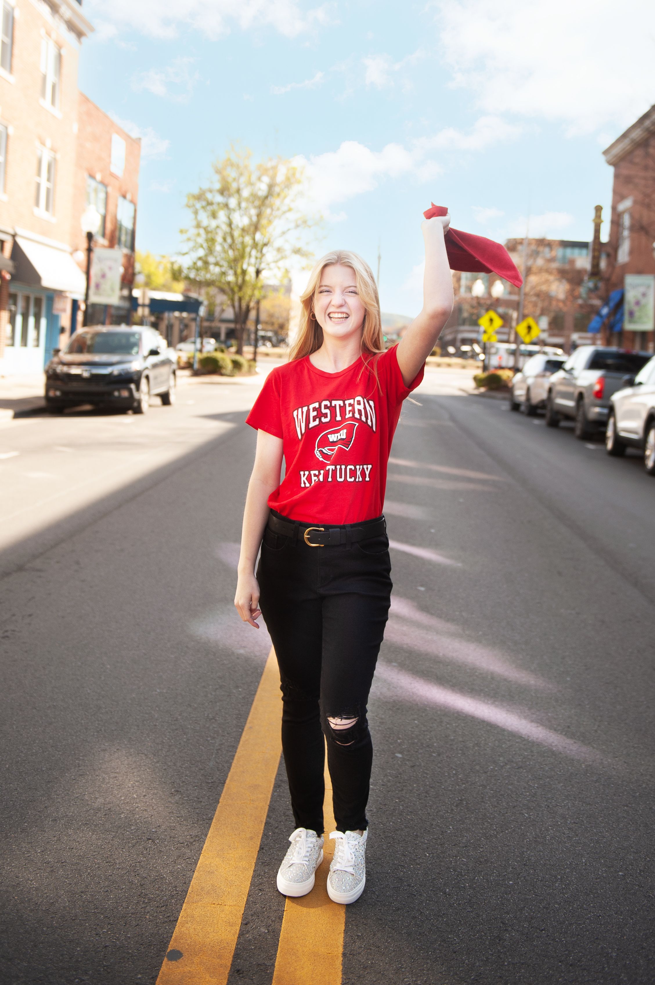 A person in a red Western Kentucky shirt and black pants stands on yellow street lines in an urban setting.