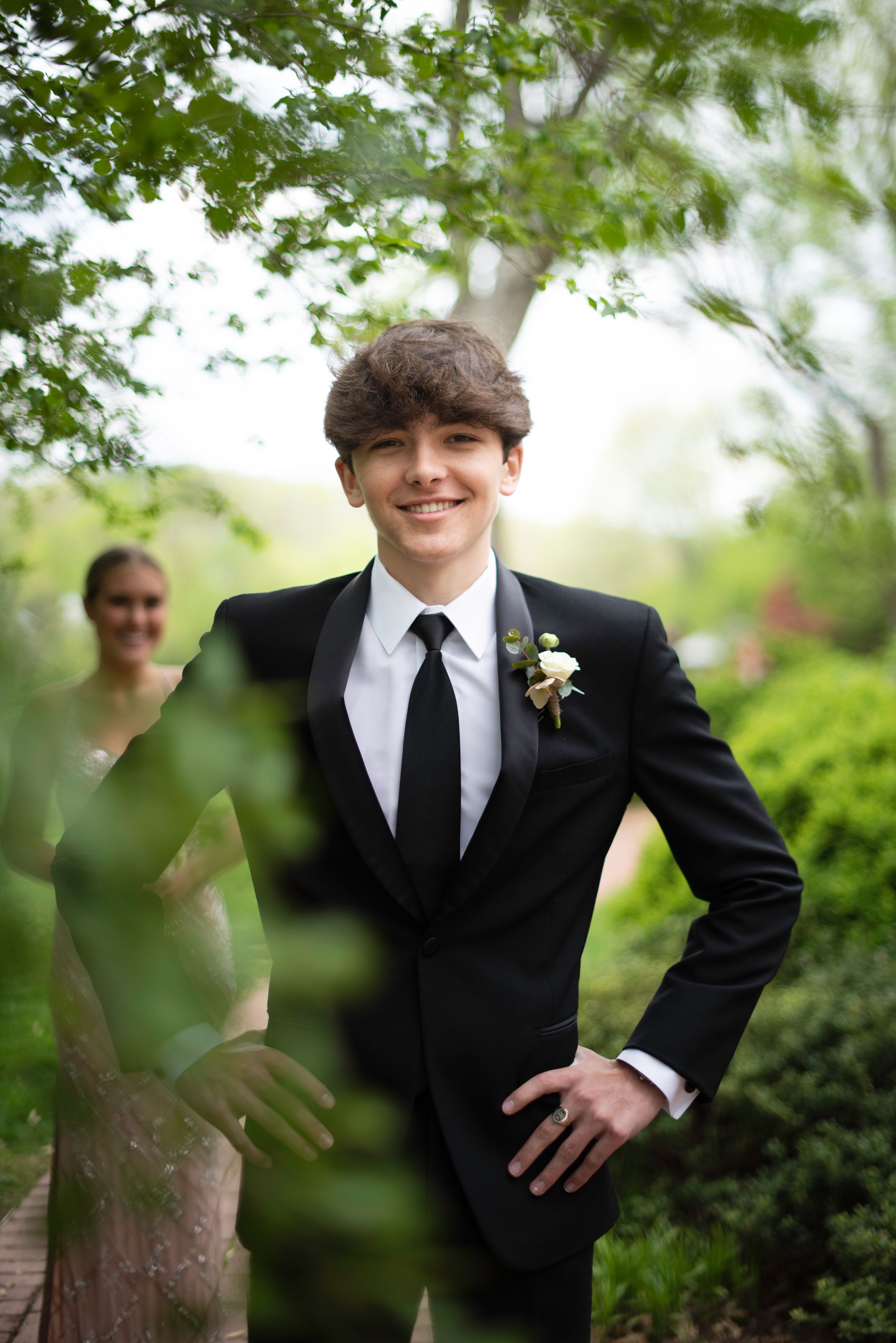 A person in formal black suit and tie stands among lush green foliage outdoors for a portrait session.
