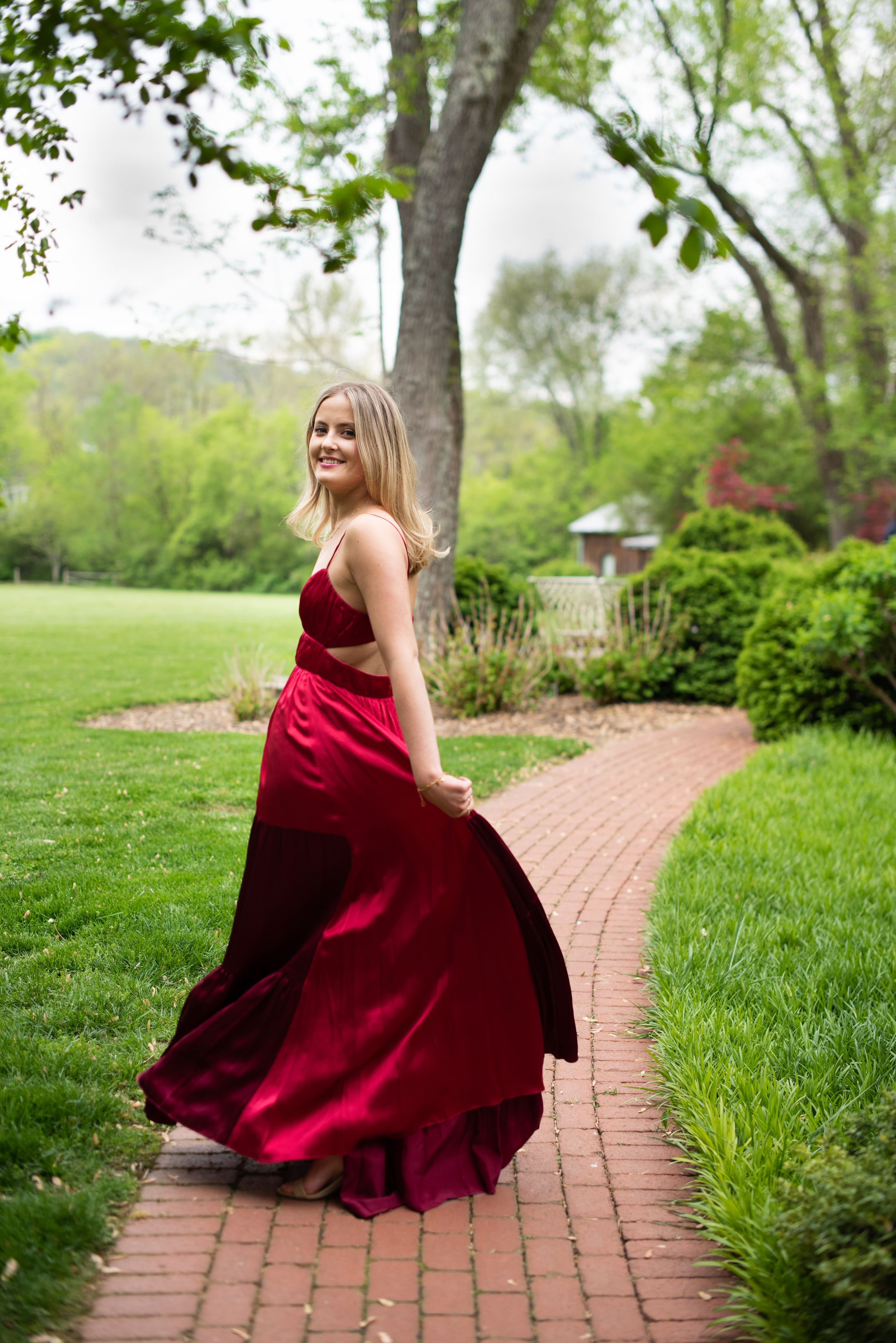 A person in a flowing burgundy formal dress twirls on a brick pathway surrounded by lush spring gardens.