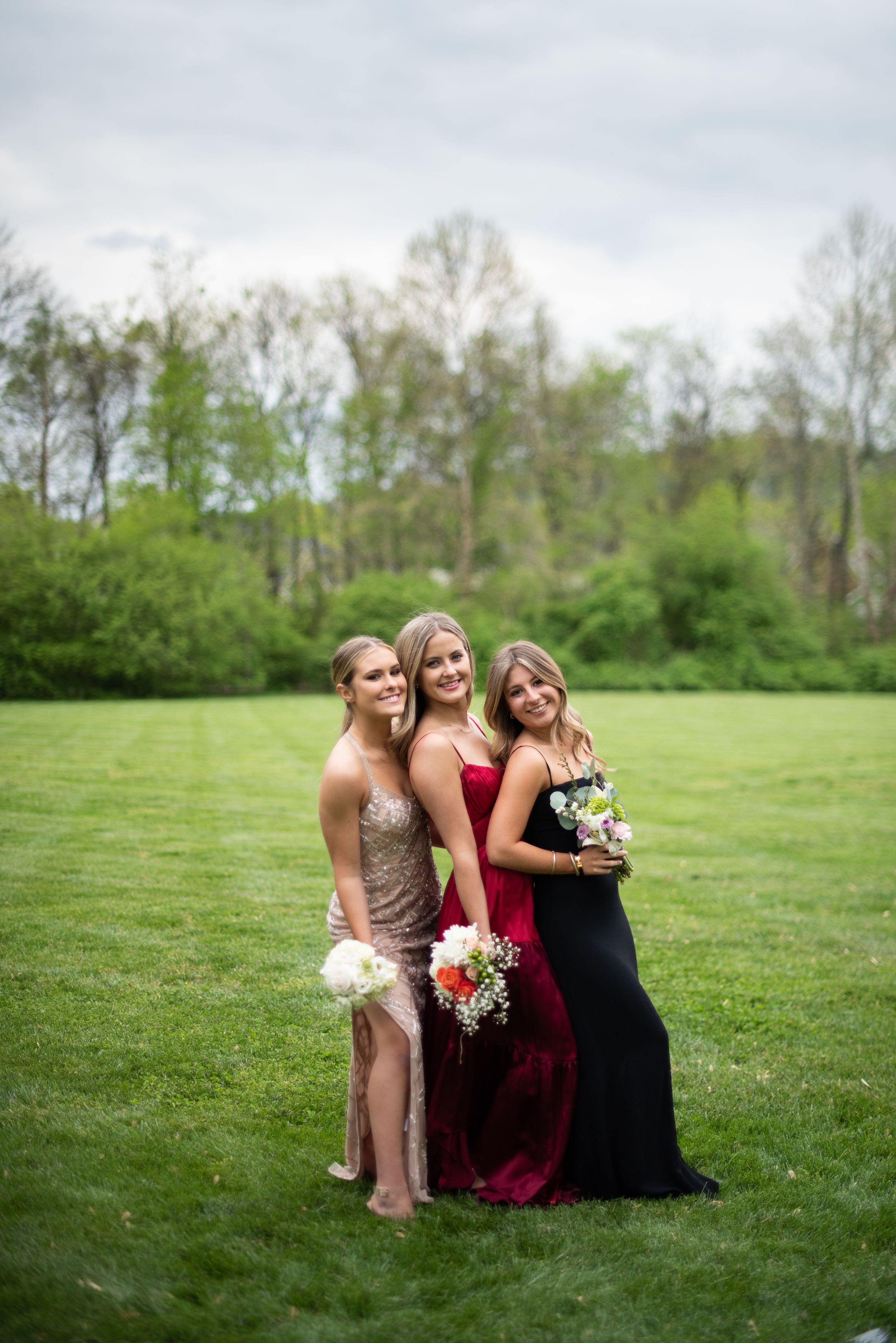 Three friends in formal dresses sit together on grass in a spring meadow with trees in the background.