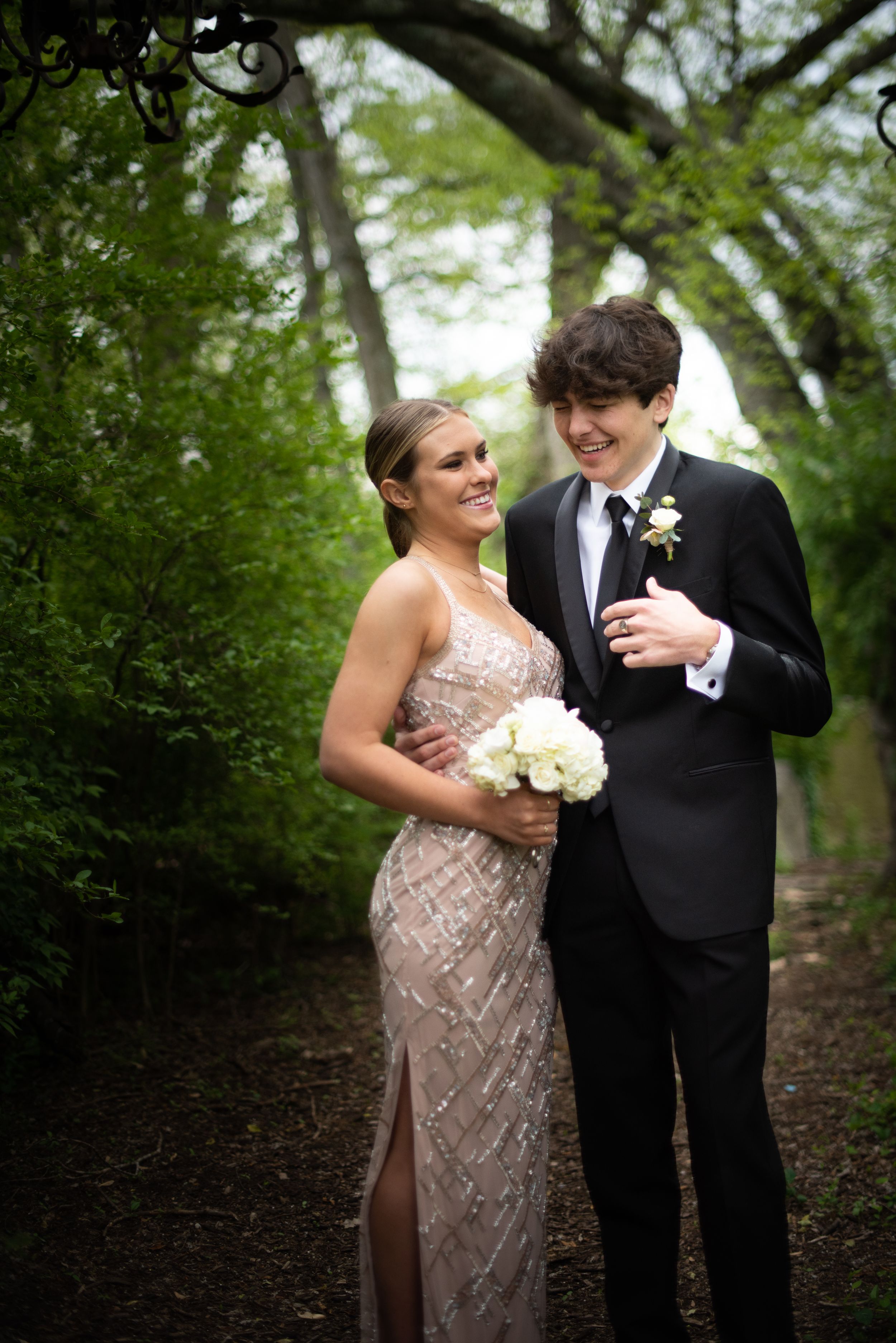 A couple in formal attire share intimate moments while walking through a lush green wooded area during their wedding photoshoot.
