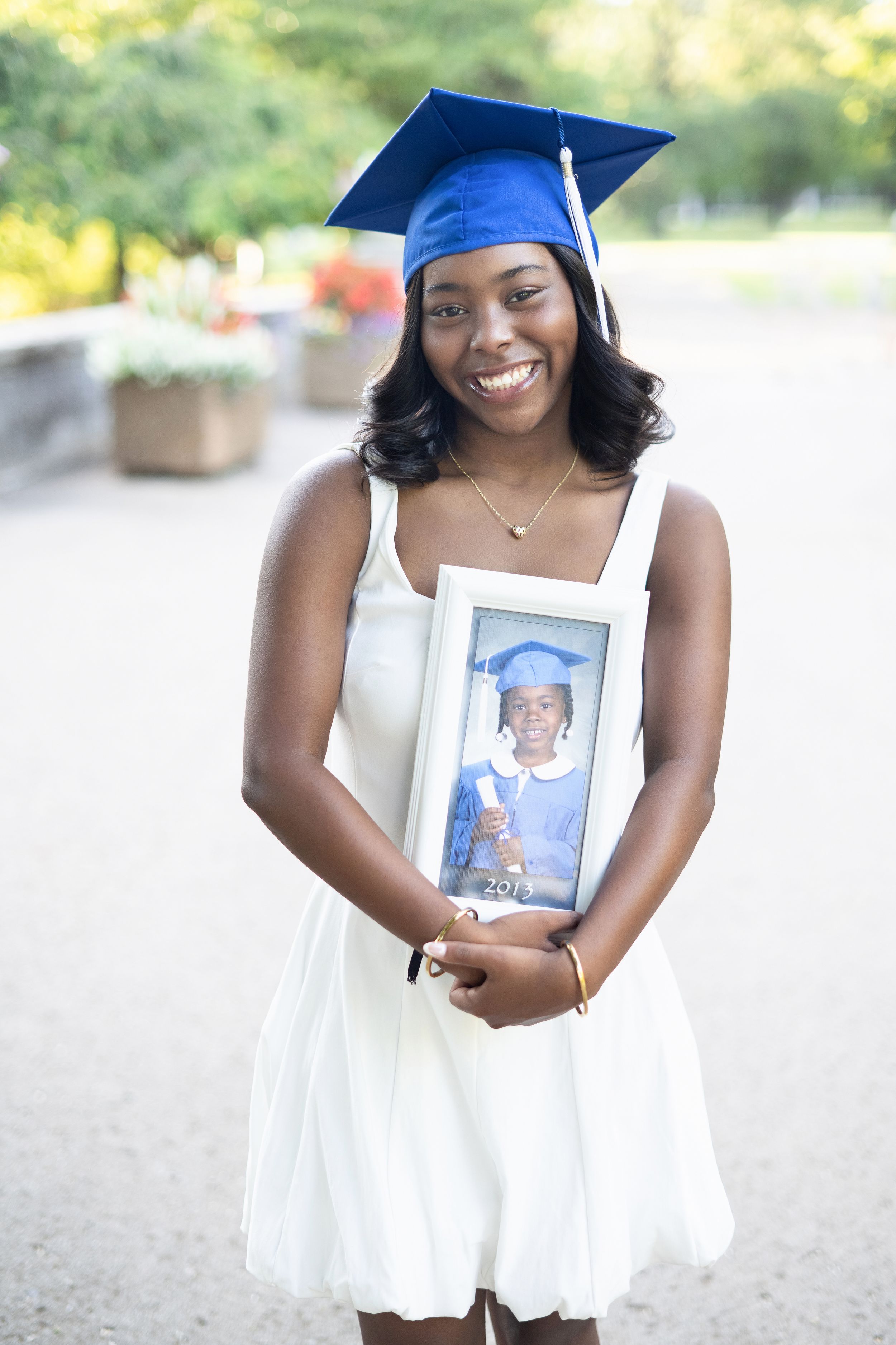 Graduate in blue cap and white dress holds framed photos during outdoor graduation photoshoot session.