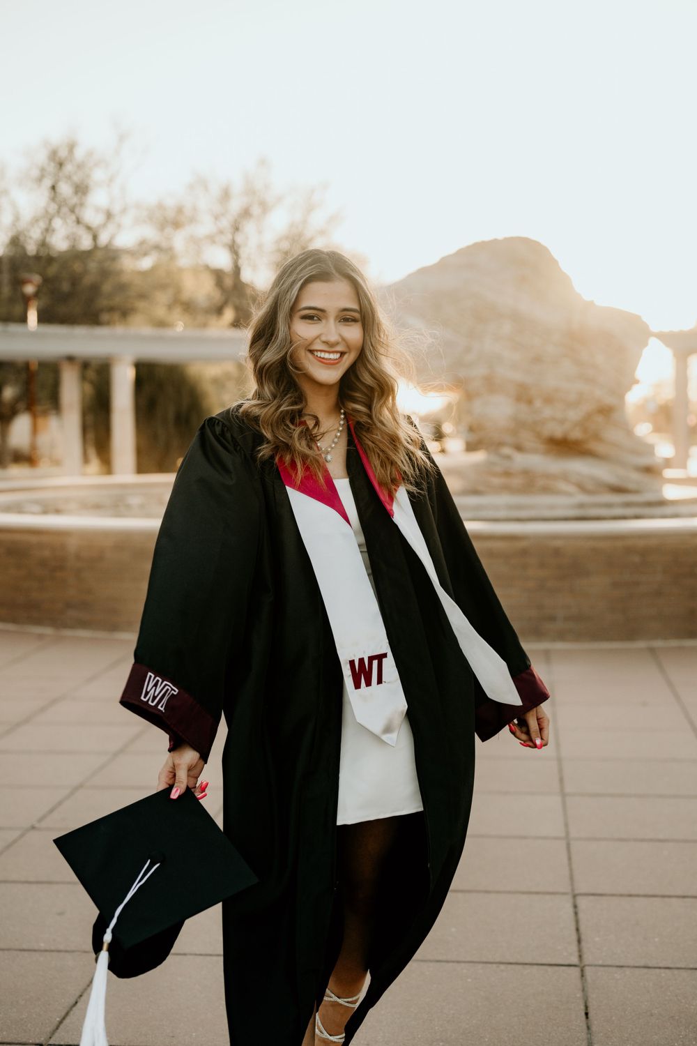 Graduate in black cap and gown celebrates during sunset campus photoshoot.