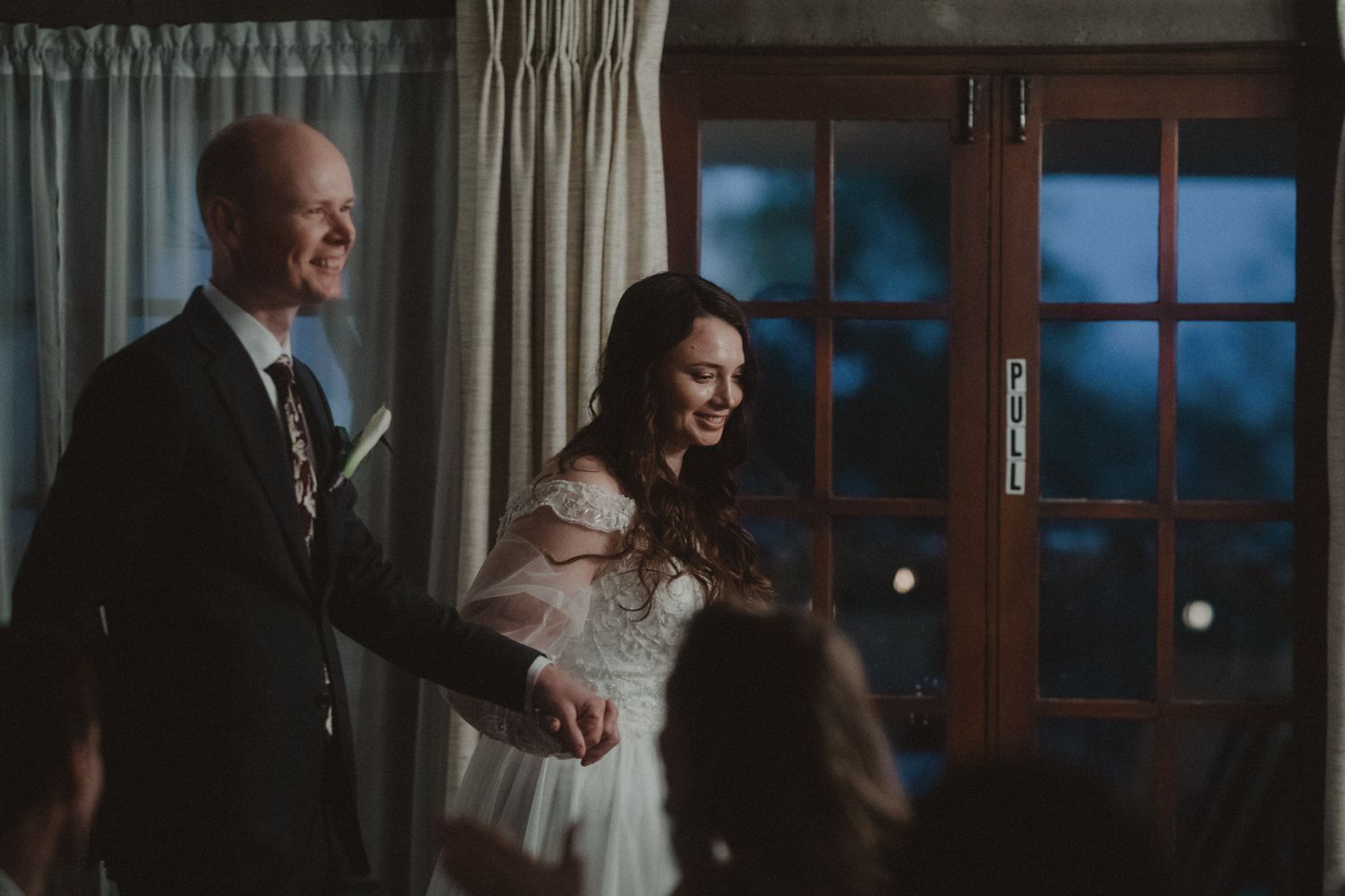 A couple shares an intimate moment while dancing together in a dimly lit room with large windows.