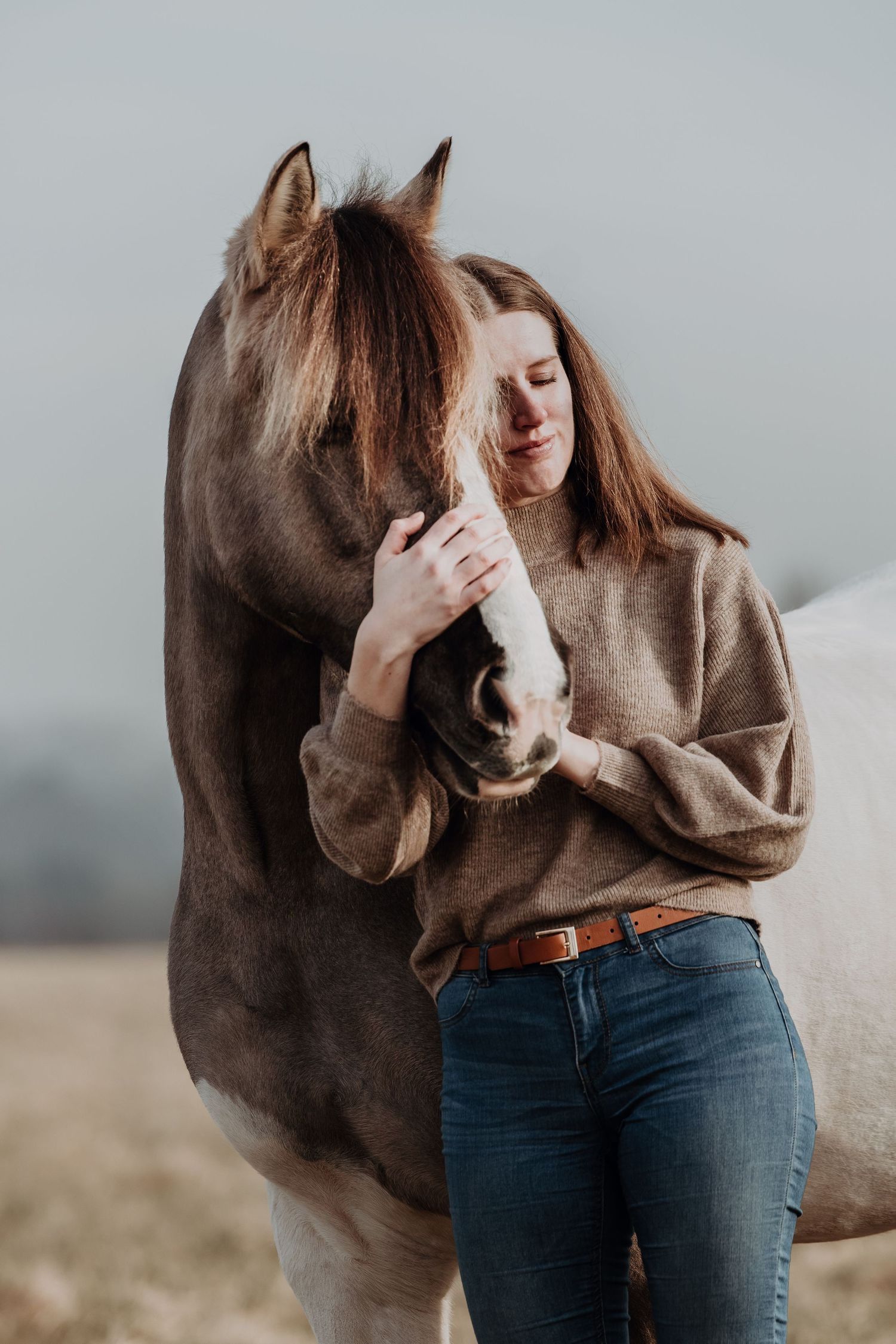 A tender moment shared between a dark brown horse and someone wearing a beige sweater and jeans in a field.