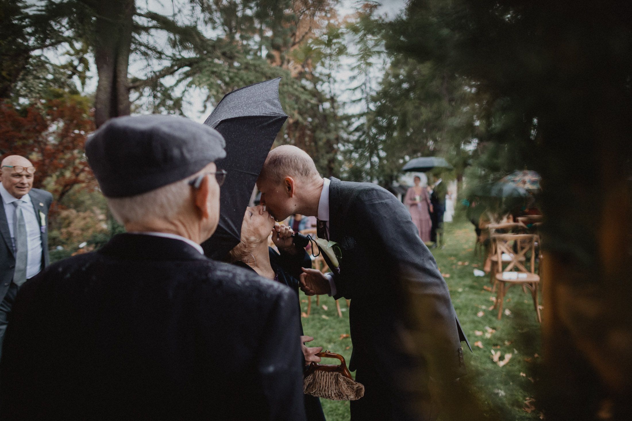 Wedding guests gather outdoors under umbrellas during a light rain at an autumn ceremony.