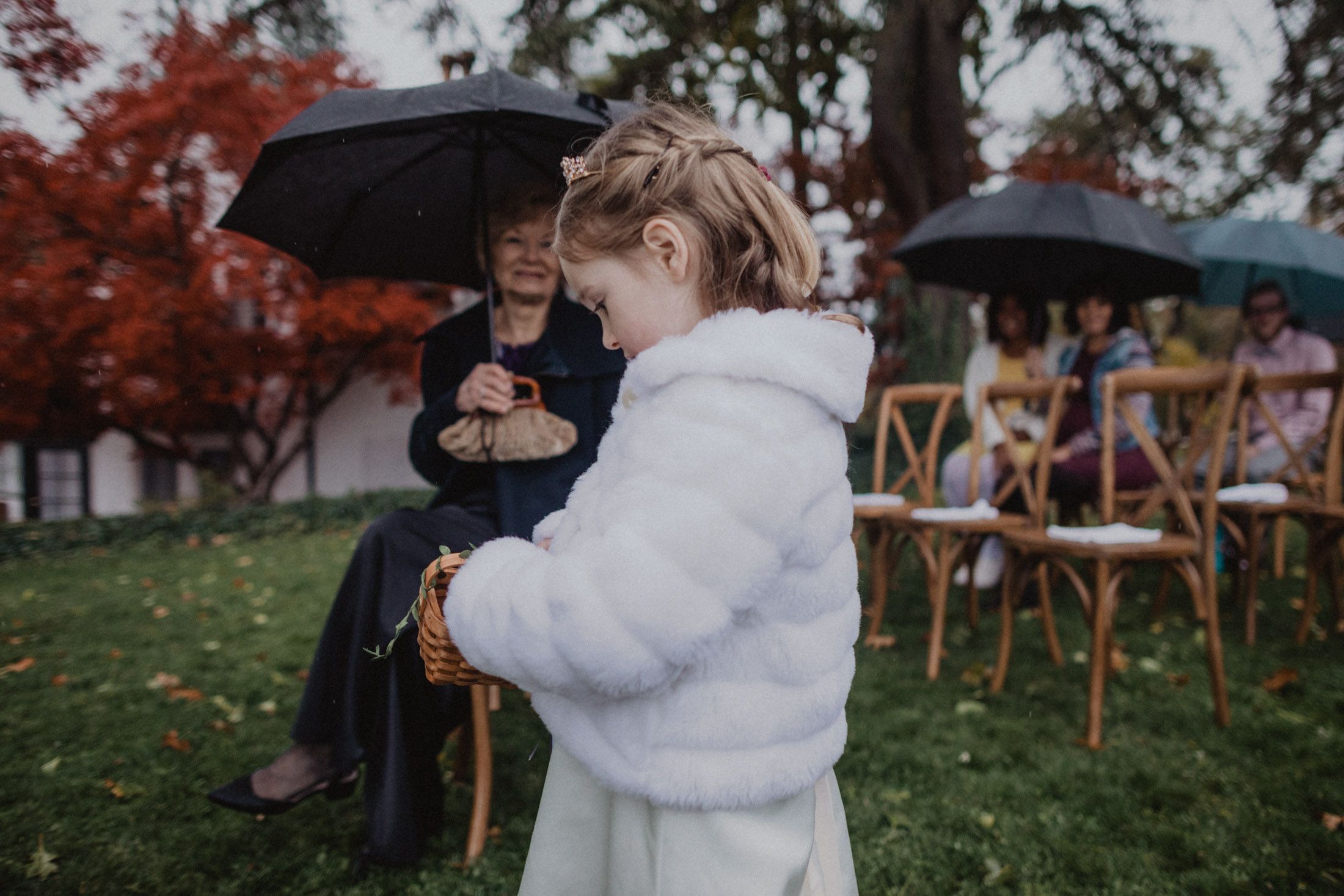 A figure in a white fur coat stands on a rainy lawn with umbrellas and wooden chairs visible in the background.