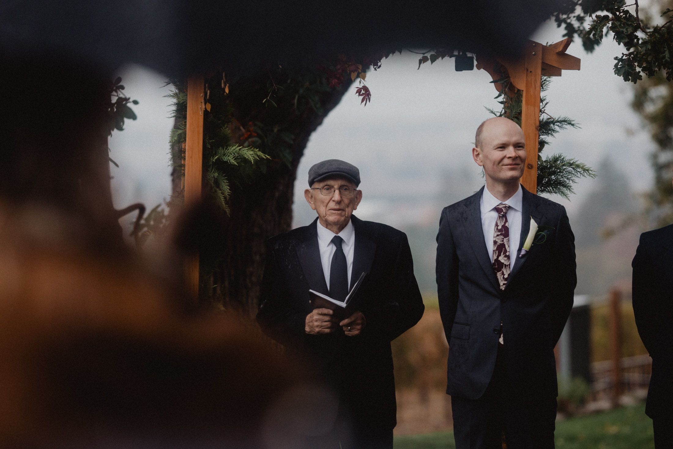 Two men in formal attire stand together at an outdoor wedding ceremony beneath an arch.