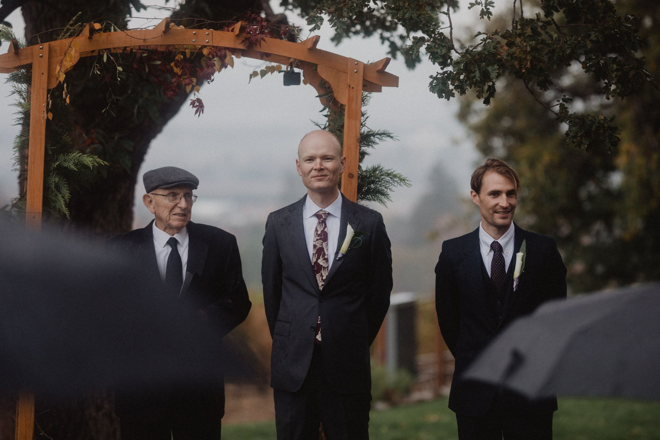 Wedding party standing under a wooden arch during an outdoor autumn ceremony.