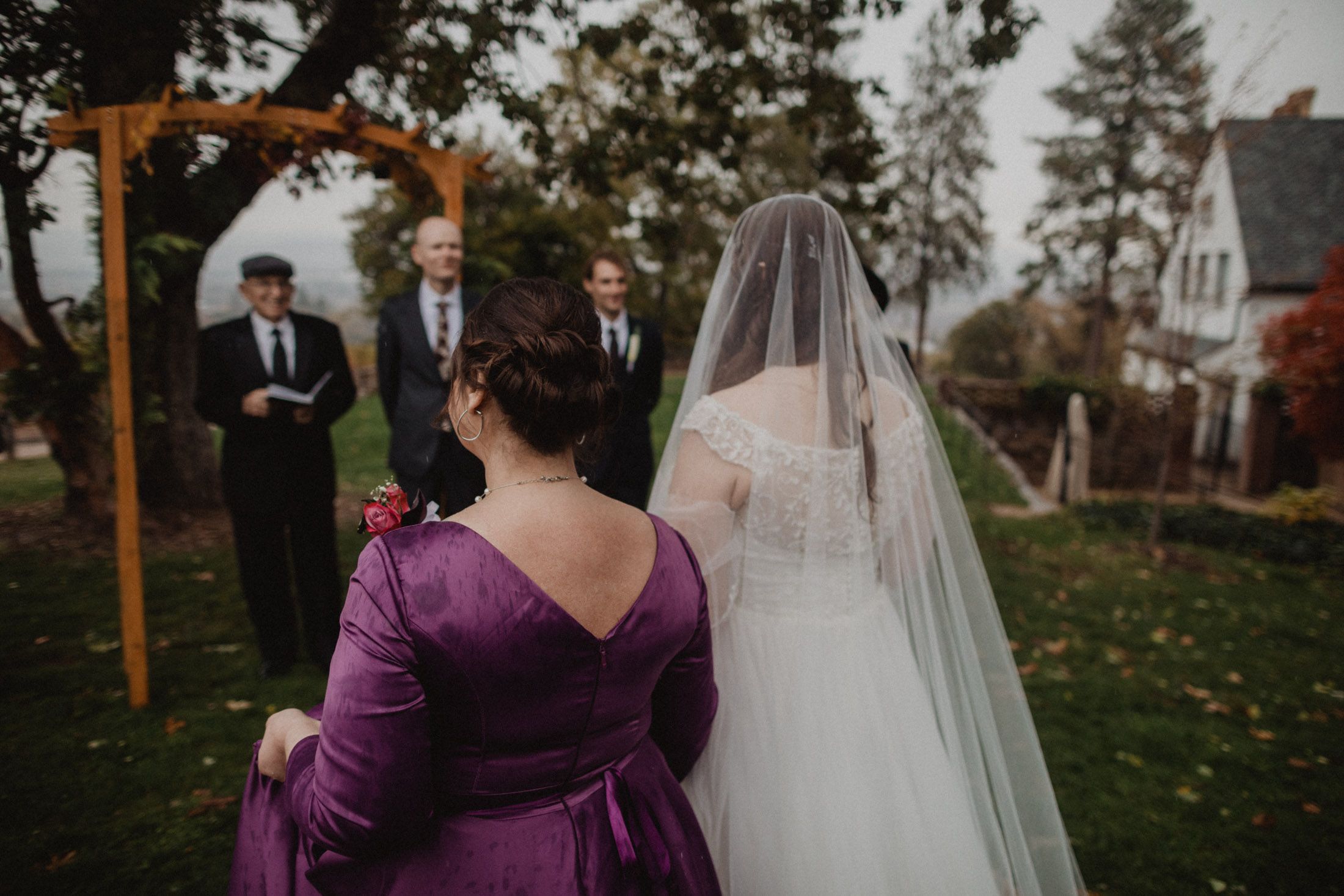 Wedding ceremony taking place outdoors with purple-clad attendants and a wooden arch.