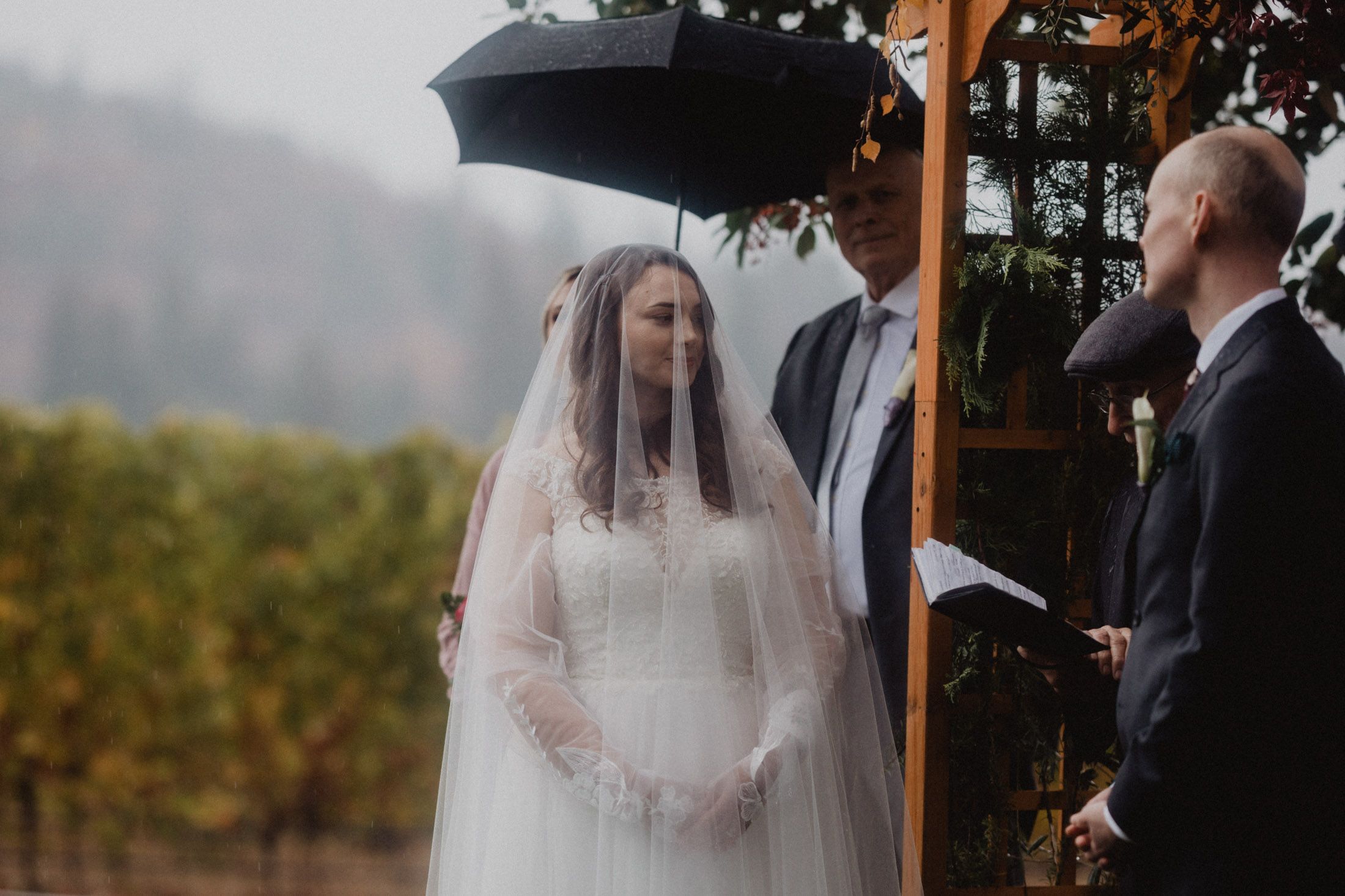 A bride in a white dress and veil stands under a black umbrella during an outdoor ceremony.