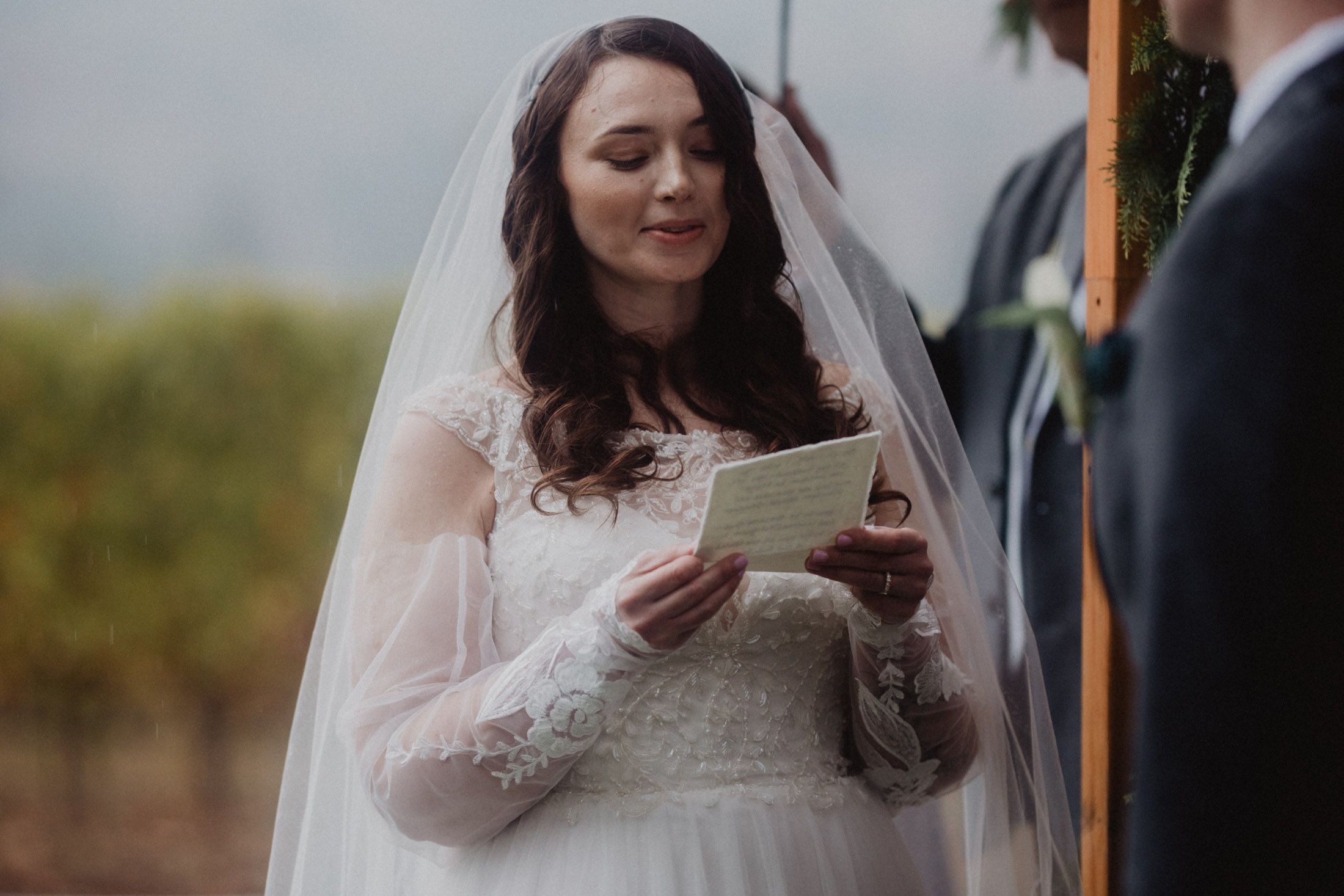A bride in a white lace wedding dress reads from a paper during an outdoor ceremony.