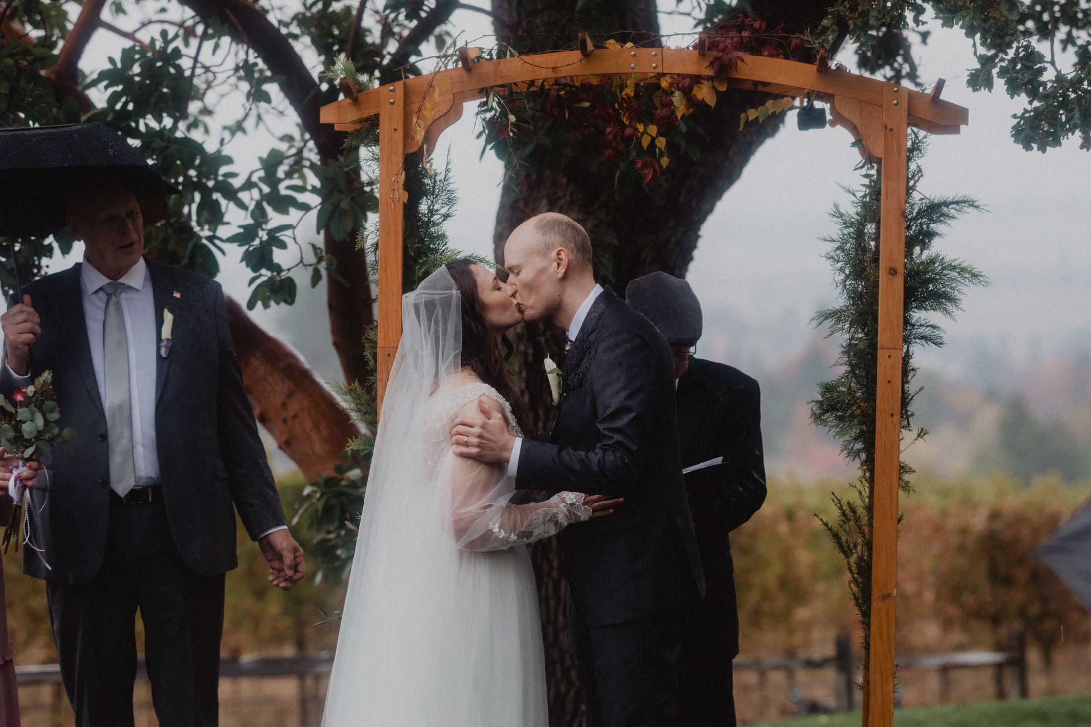 A couple stands under a wooden pergola during an autumn wedding ceremony surrounded by guests and fall foliage.