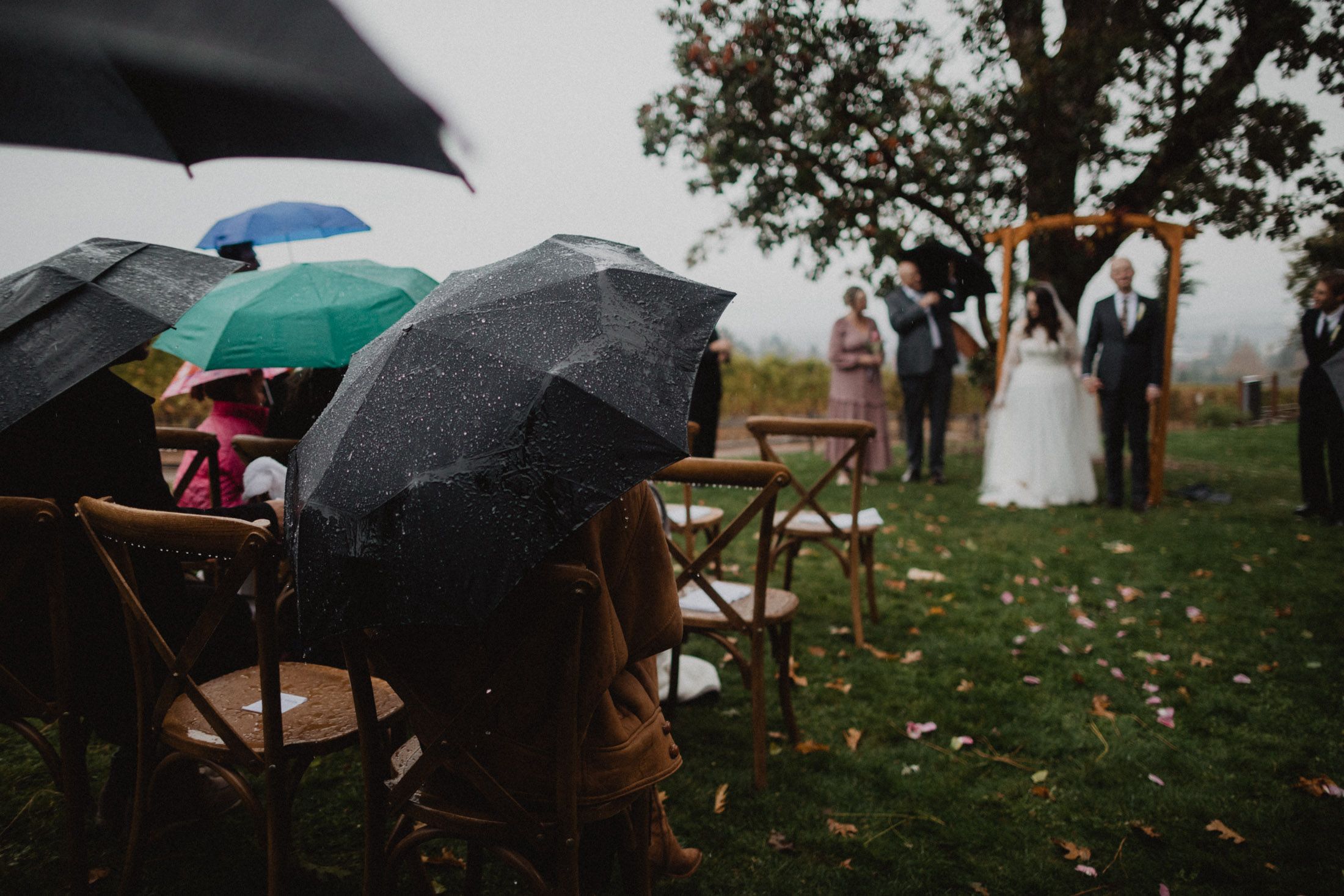 Umbrellas shield wedding guests during a rainy outdoor ceremony with autumn leaves scattered on the ground.