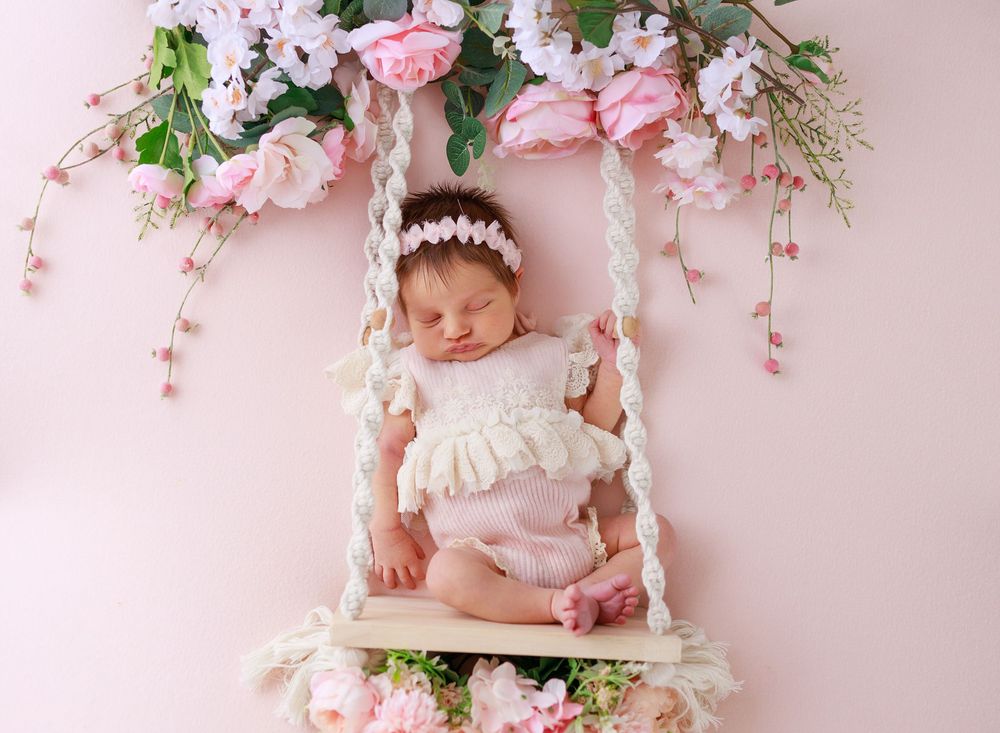 Newborn sleeping on decorative swing adorned with pink flowers and floral arrangements.