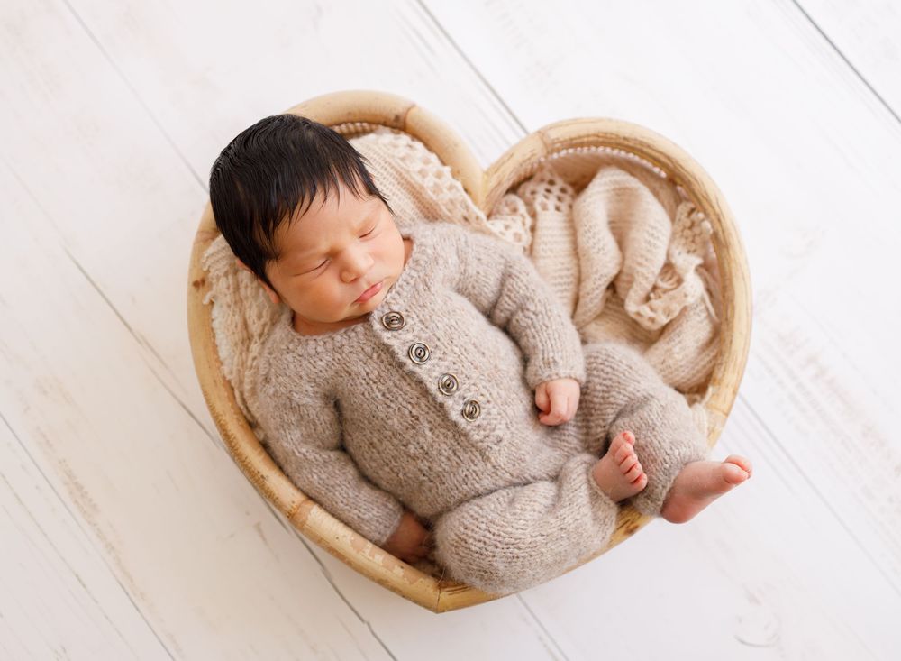 Sleeping infant wearing knit outfit nestled in heart-shaped basket on white wooden floor.