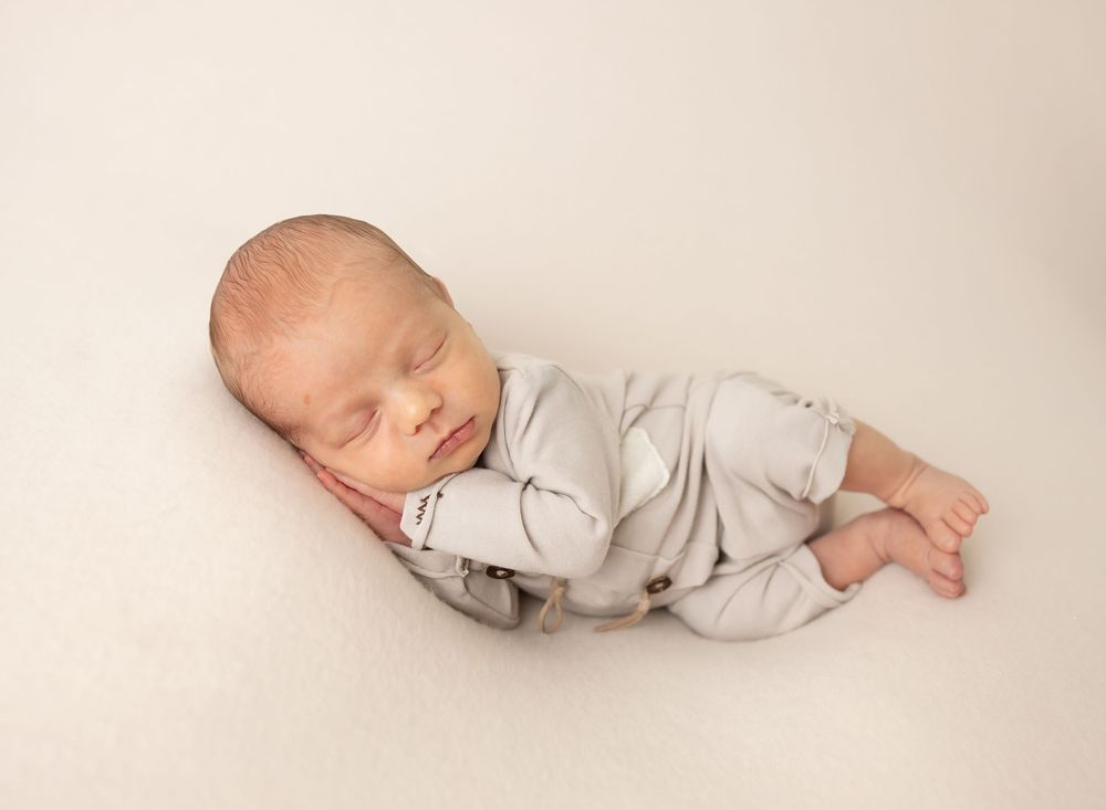 Peaceful sleeping newborn wearing light gray outfit on neutral backdrop.