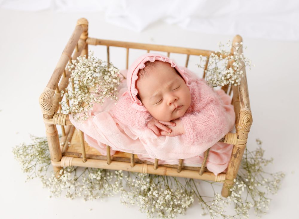 Sleeping baby in pink outfit resting in wooden doll bed surrounded by baby's breath flowers.