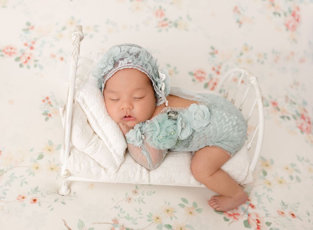 Sleeping baby in vintage-style mint green outfit on white iron bed with floral backdrop.