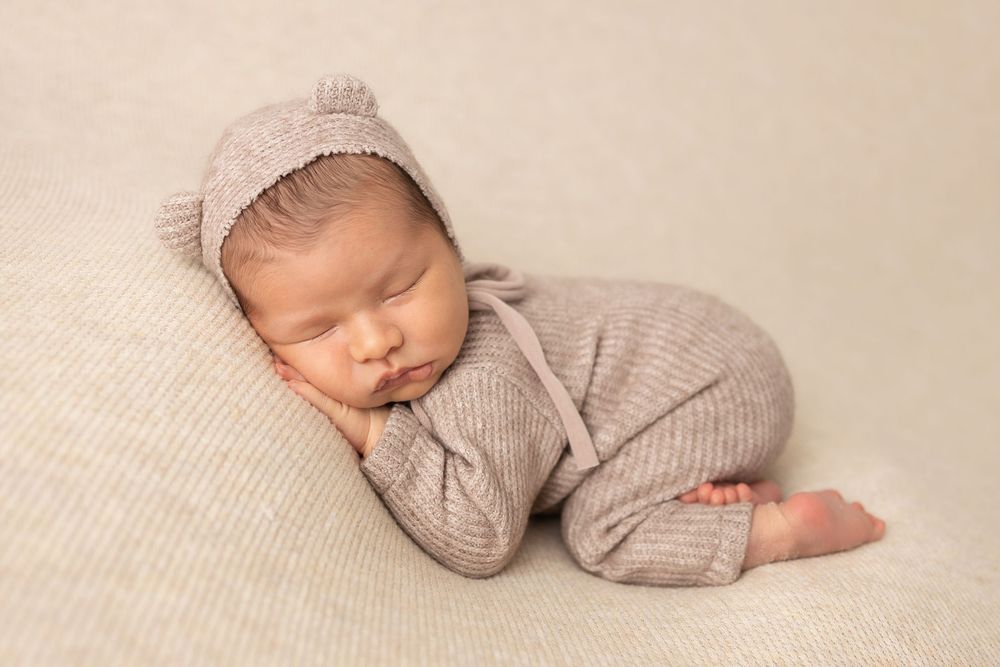 Sleeping infant in knit beanie and sweater posed on textured beige backdrop.