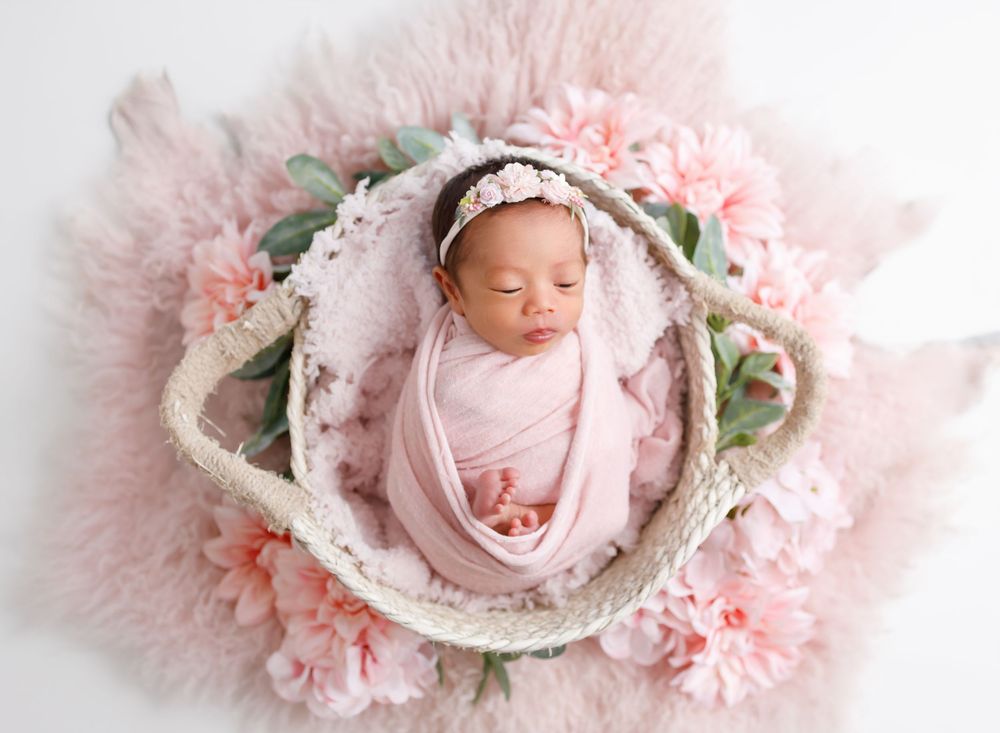 Sleeping newborn in pink wrap nestled in floral basket with pink peony decorations.