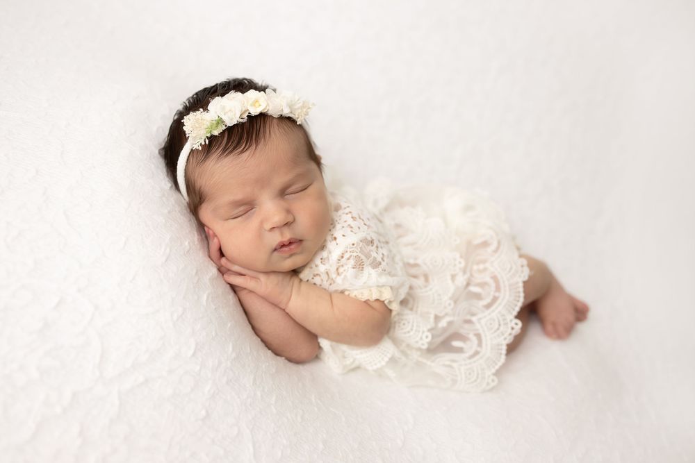 Peaceful newborn wearing white lace dress resting on soft white backdrop.