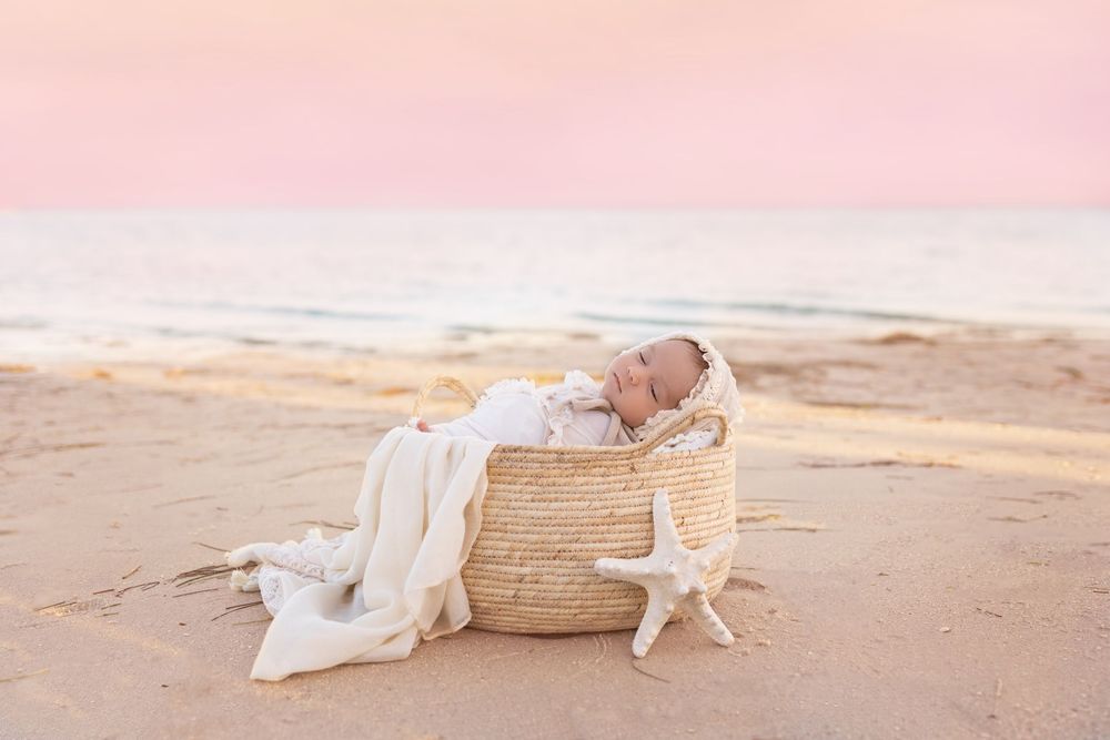 Newborn sleeping peacefully in woven basket on beach at sunset with starfish decoration.