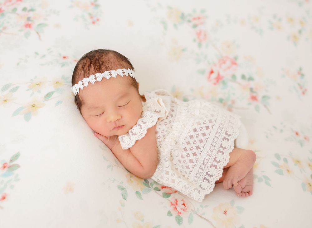 Sleeping newborn in white crochet outfit with pearl headband on floral backdrop.