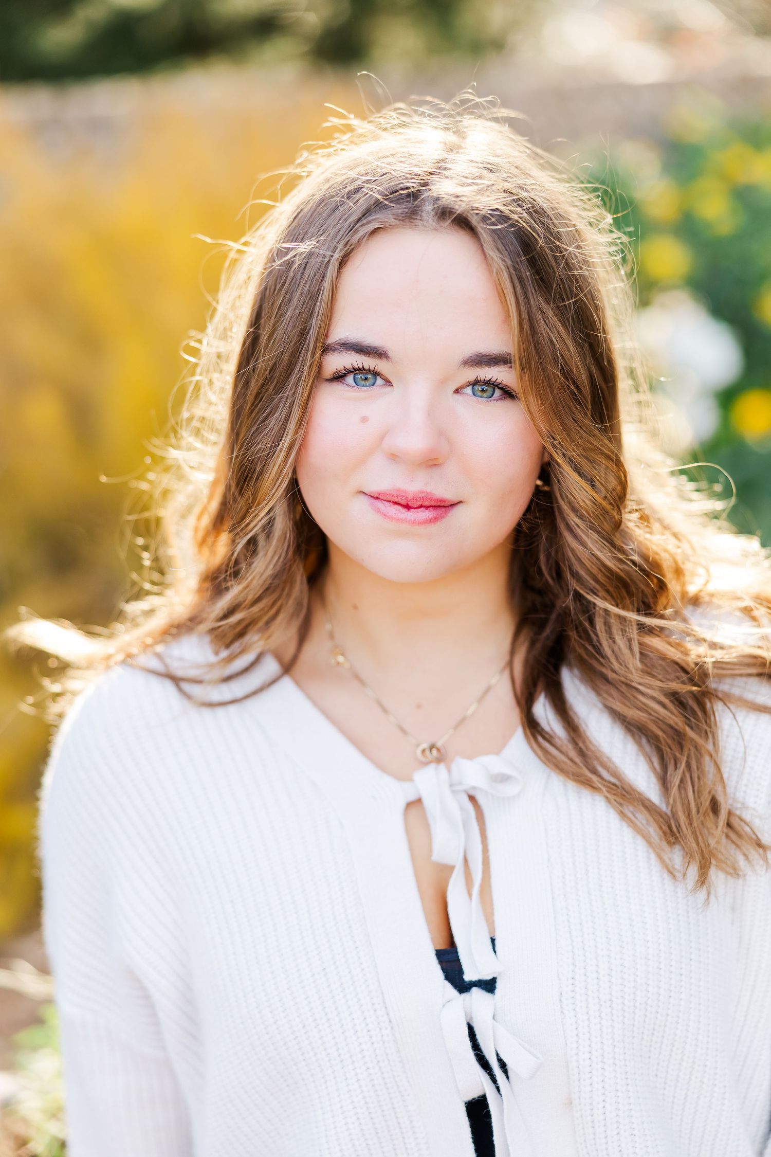 A young woman in a white sweater smiles outdoors on a sunny day with a blurred nature background at her senior photo session