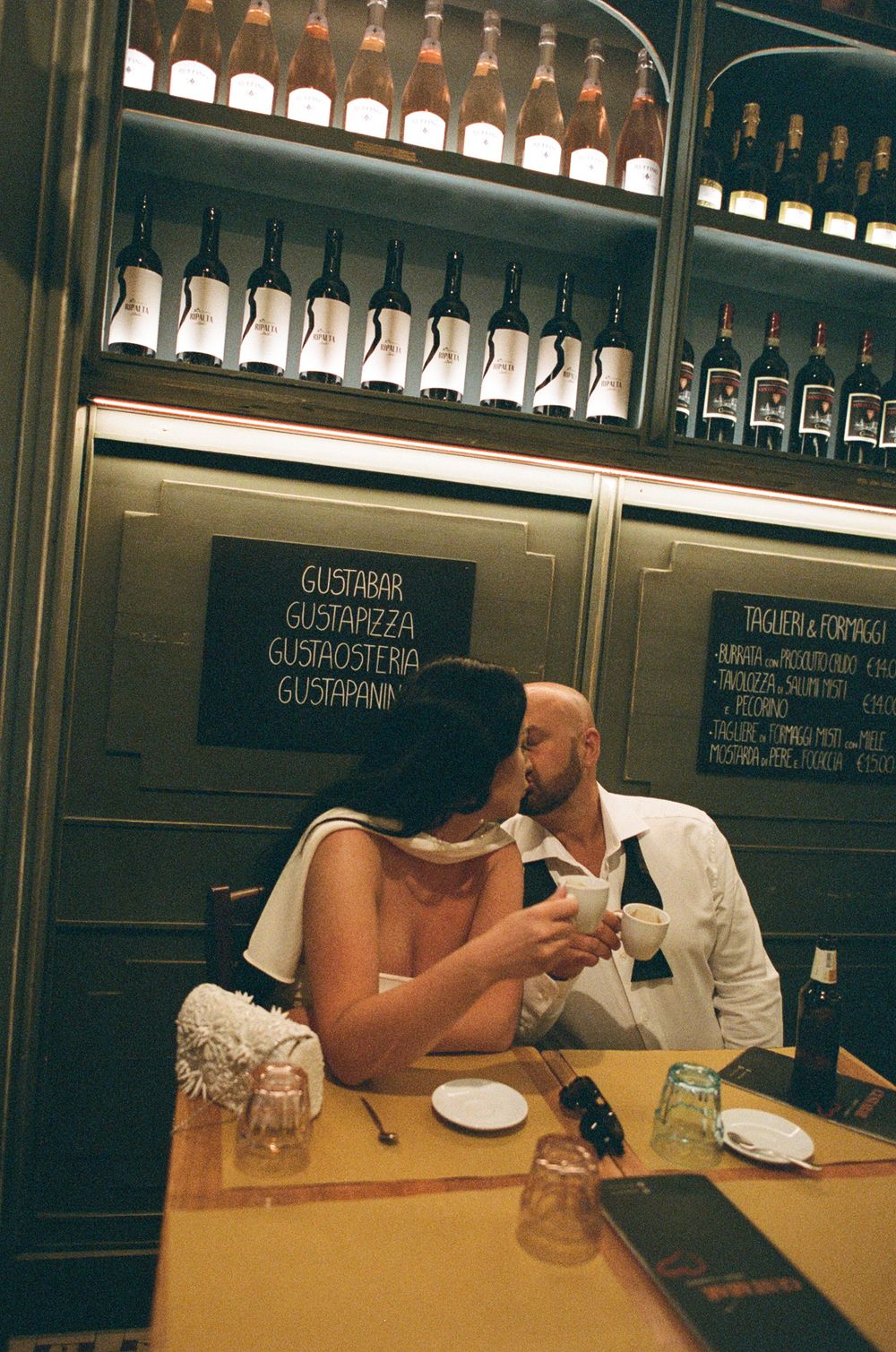 Couple sharing intimate moments at a bar with wine bottles displayed on shelves in the background.