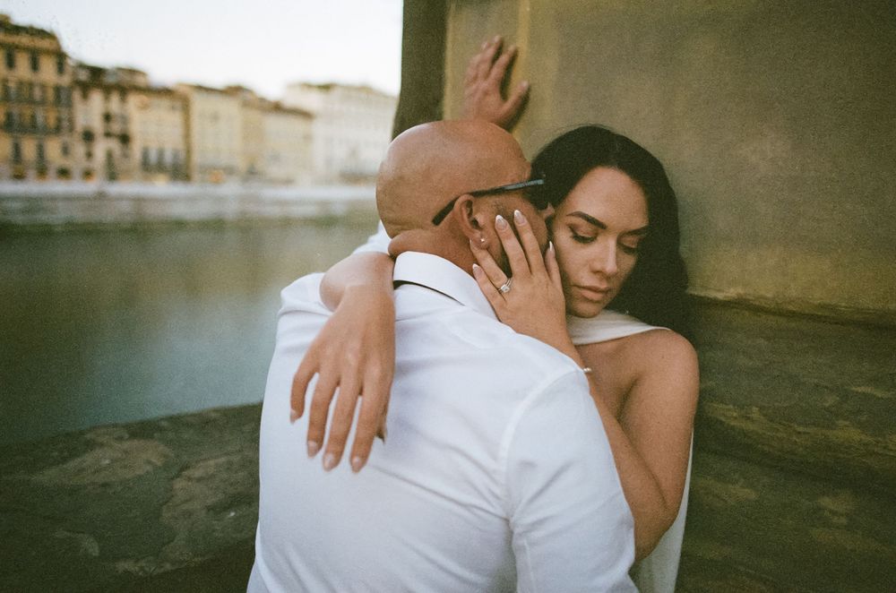 A tender embrace between two people against a backdrop of historic Italian architecture and waterfront.