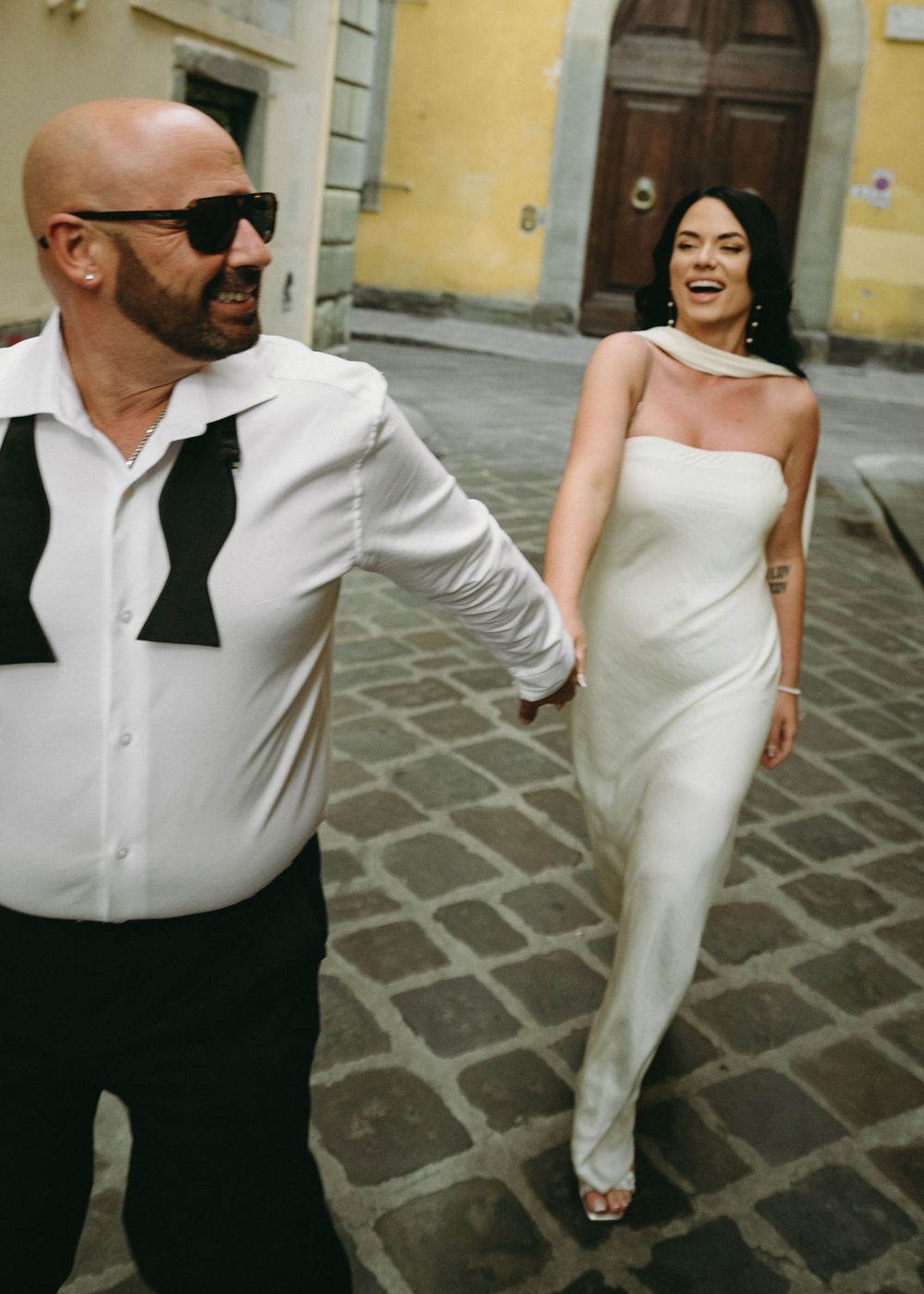 A couple in formal wedding attire dancing and laughing together on a stone paved courtyard.