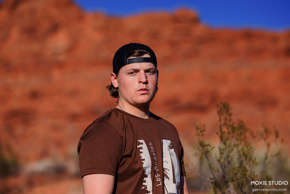 A person in a brown t-shirt and backwards cap poses against vibrant red rock formations under a bright blue desert sky.