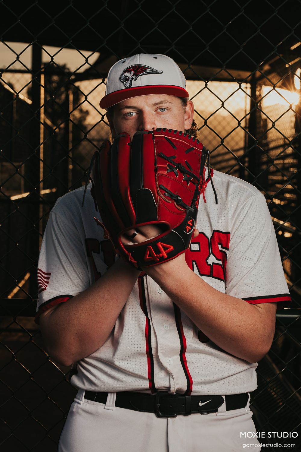 Baseball player in white uniform with red trim stands against chain link fence at sunset with red catcher's mask.