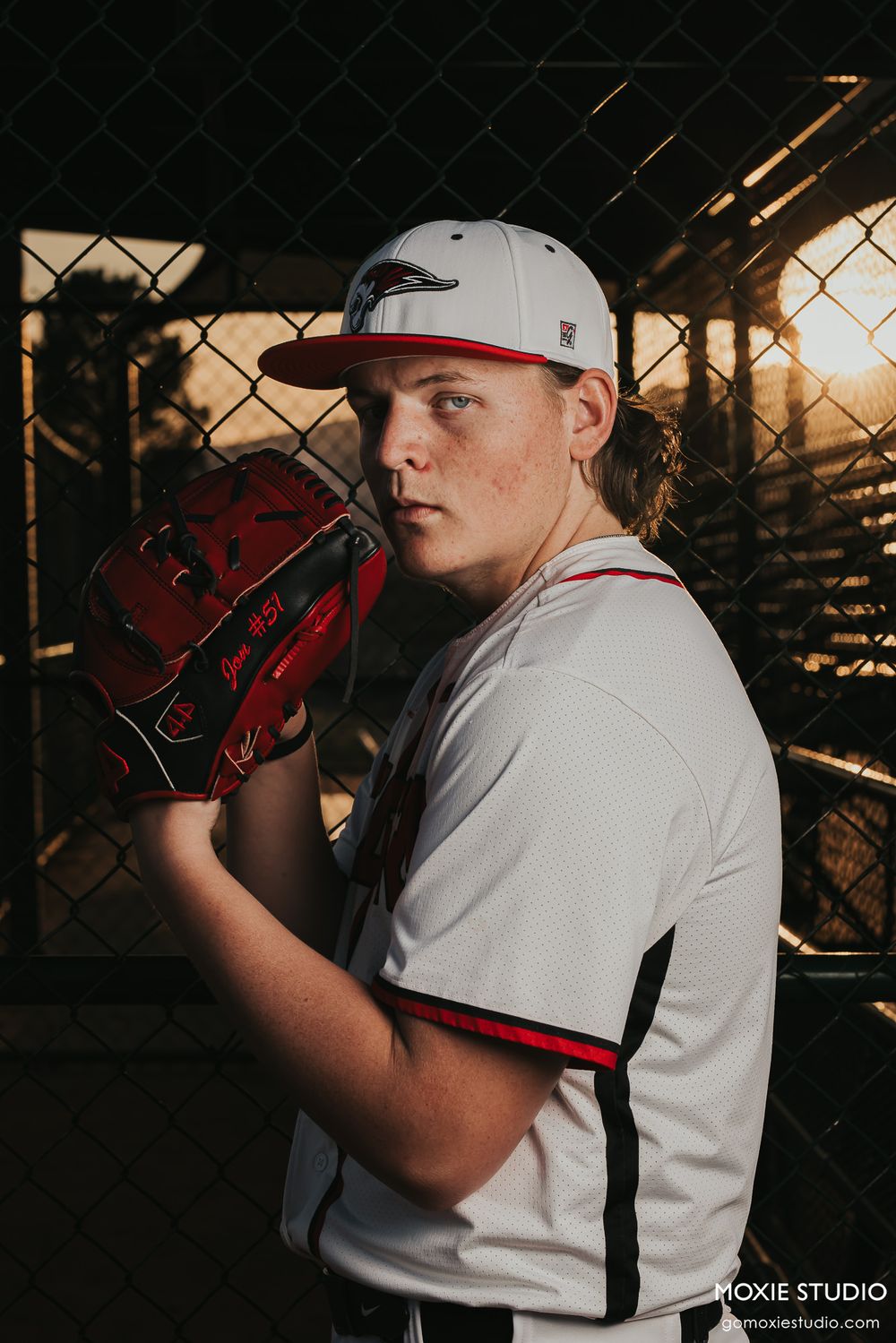 Baseball player in white uniform poses with glove at sunset behind chain link fence.