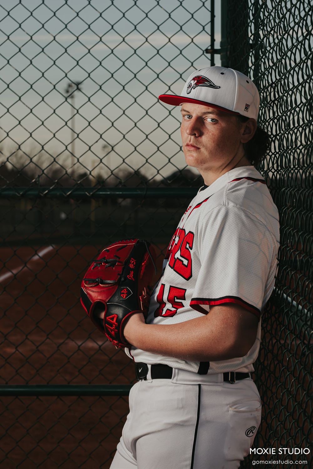 Young baseball player in white uniform and red hat poses with glove against chain link fence during practice session.