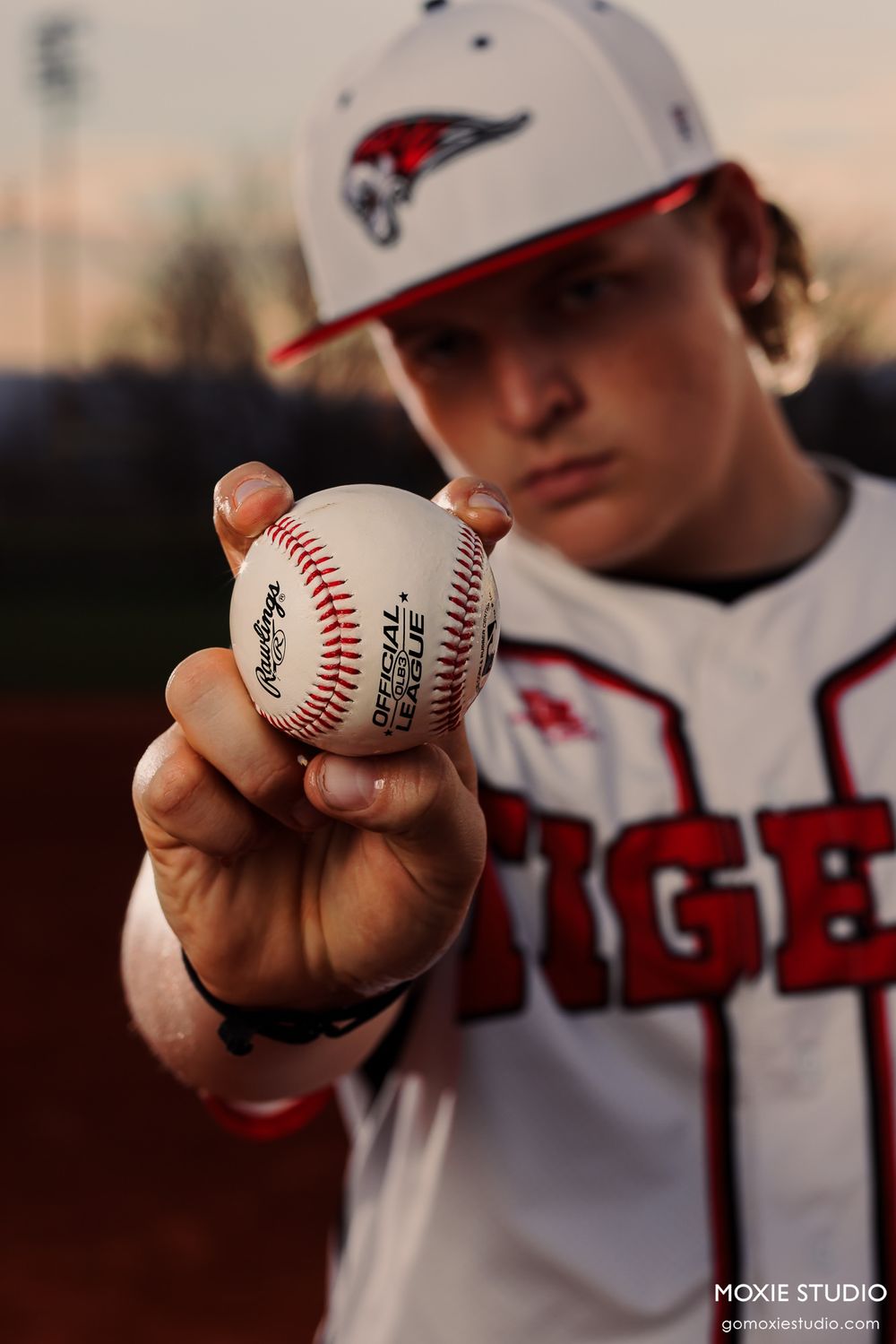 Baseball player in white uniform and cap holds up an official baseball against sunset backdrop.