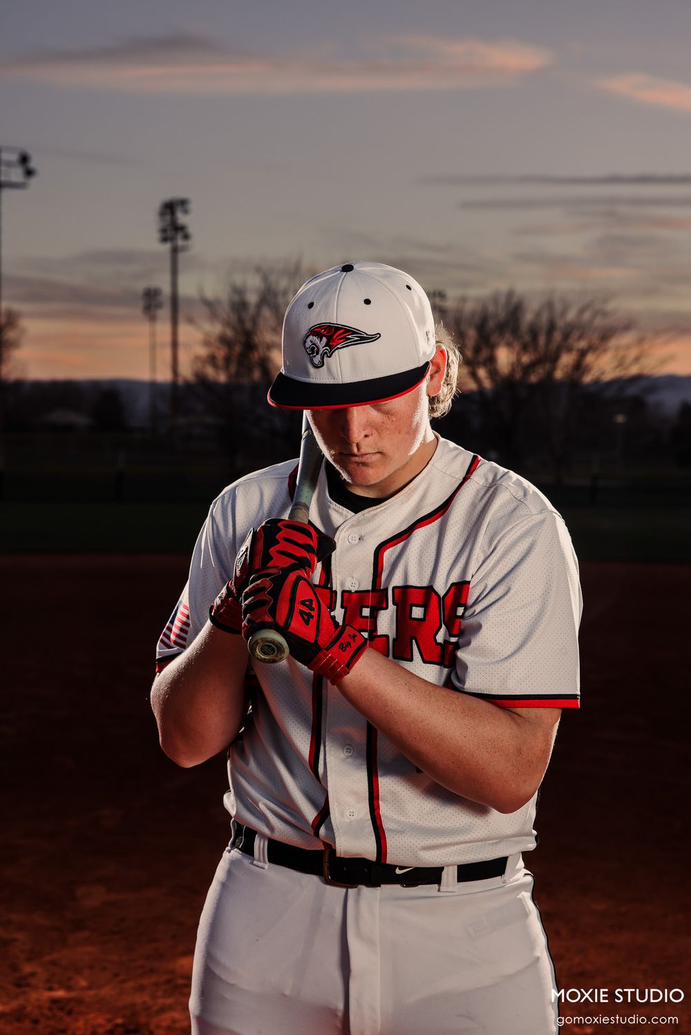 Baseball player in white uniform and cap practices batting stance at sunset during training session.