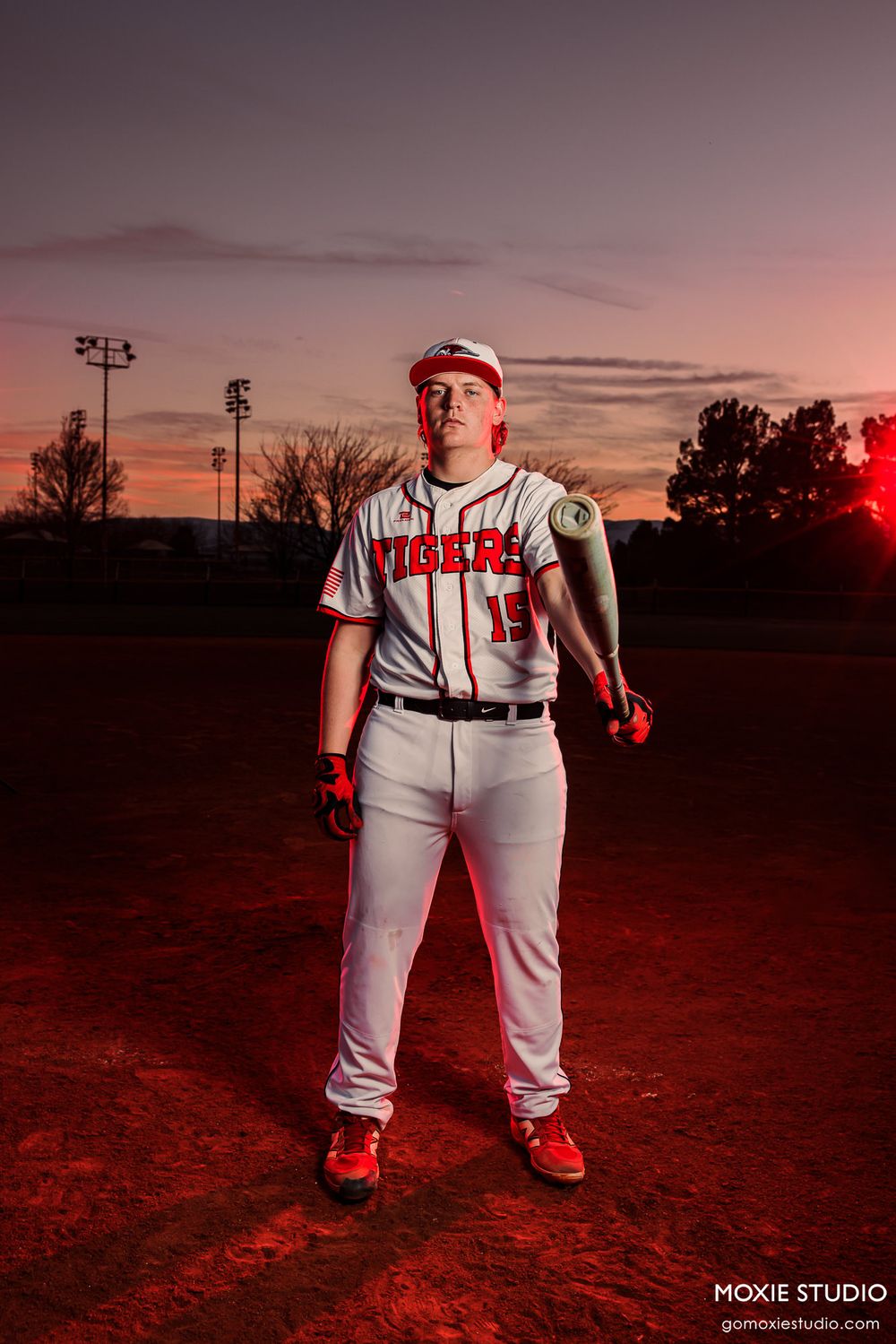 Baseball player in white uniform stands on field at sunset with dramatic red lighting and stadium lights in background.