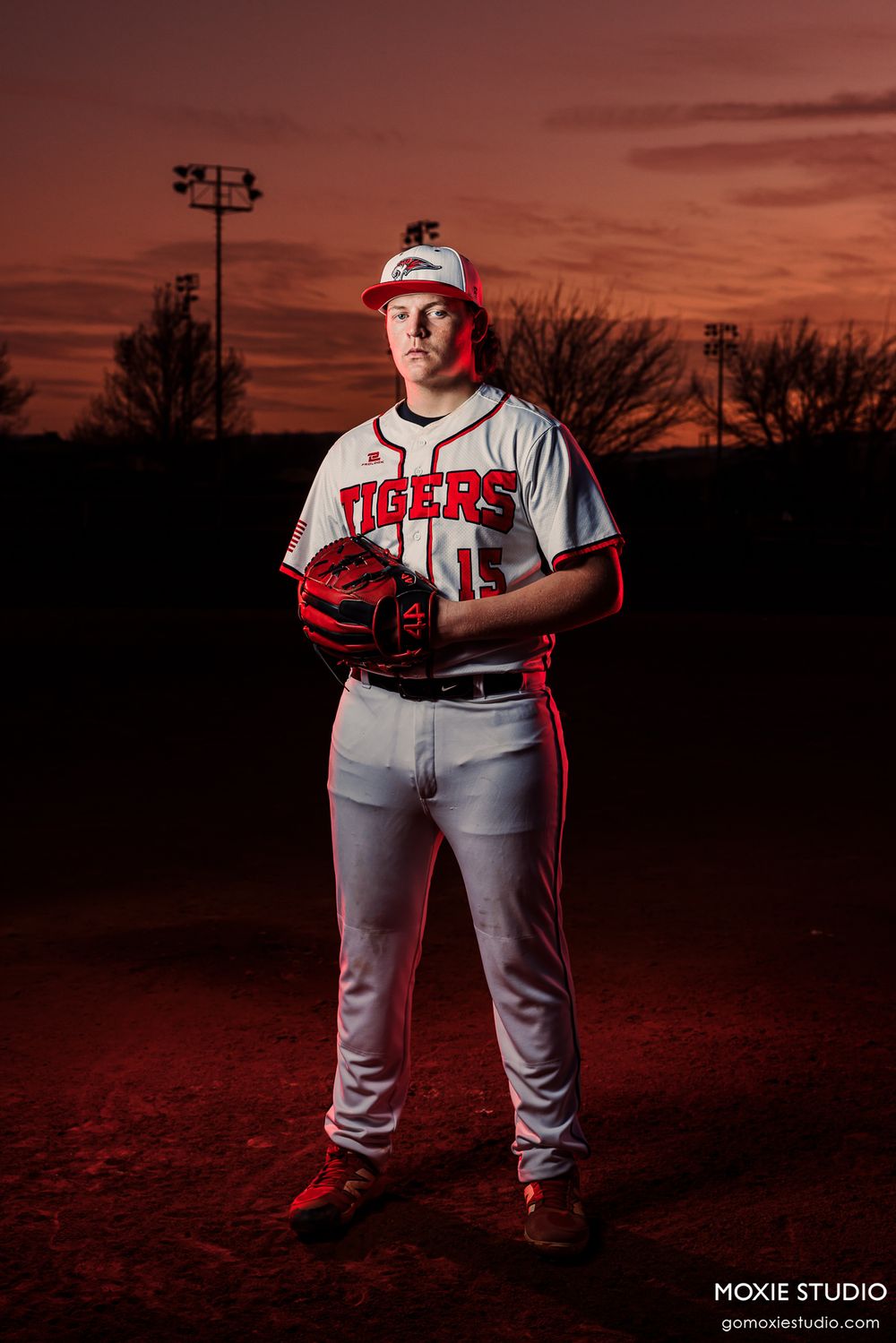 Baseball player poses in Tigers uniform against dramatic red sunset with stadium lights in background.