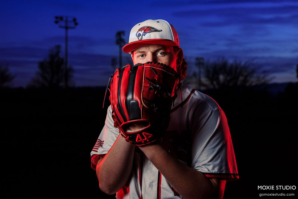 Baseball player in dramatic blue lighting poses with glove covering face on dark field at dusk.