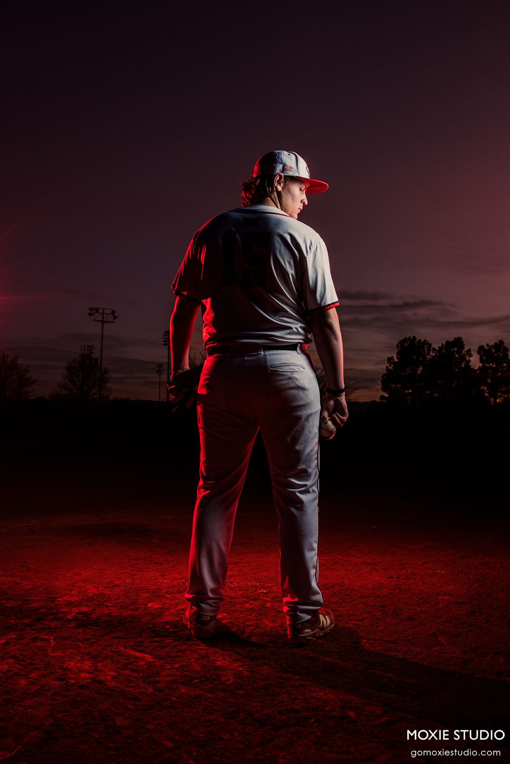 Silhouetted figure stands in dramatic red-lit field against dark sky with power lines visible in background at dusk.