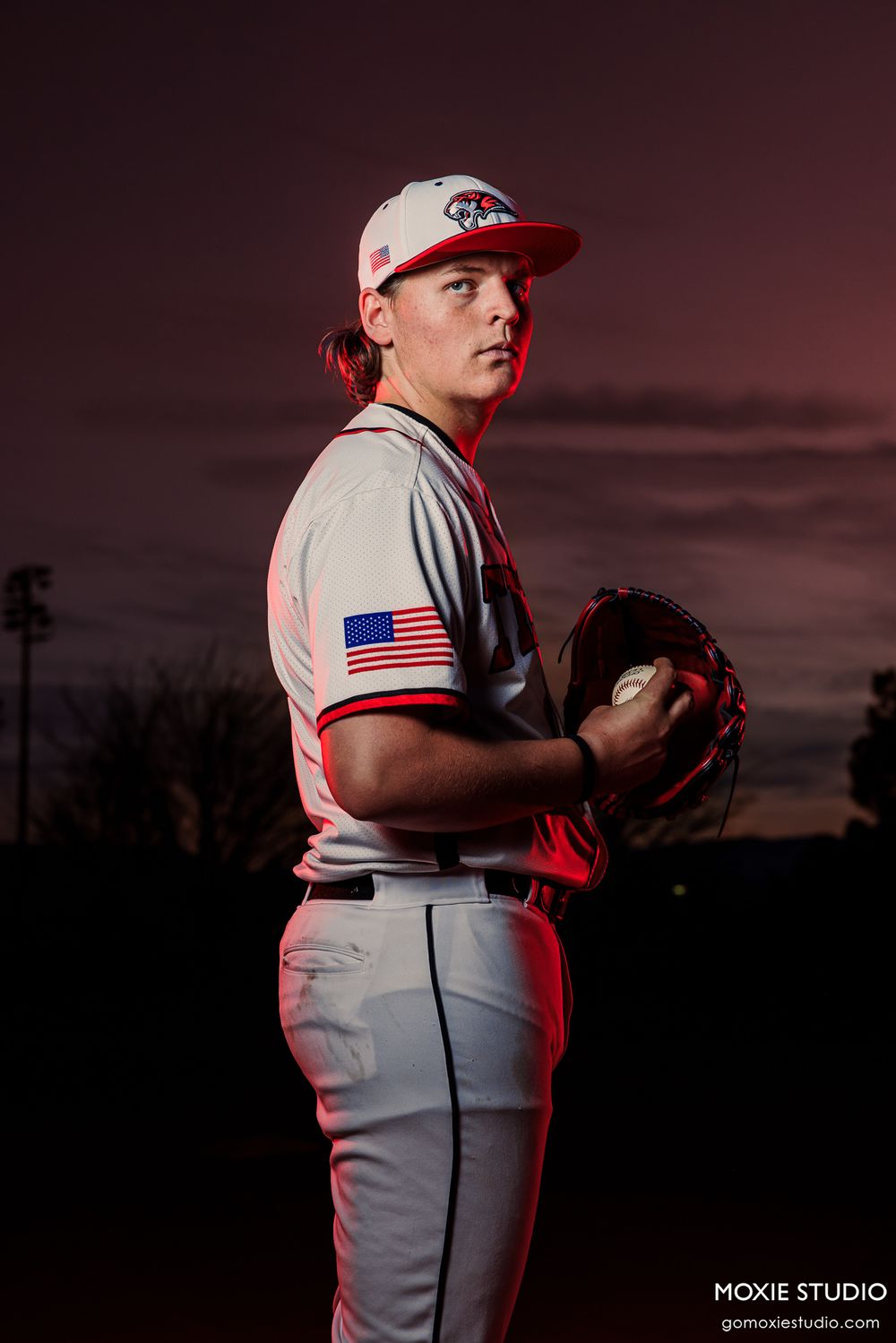 Baseball player in grey uniform with American flag patch stands ready in pitching stance at dusk.