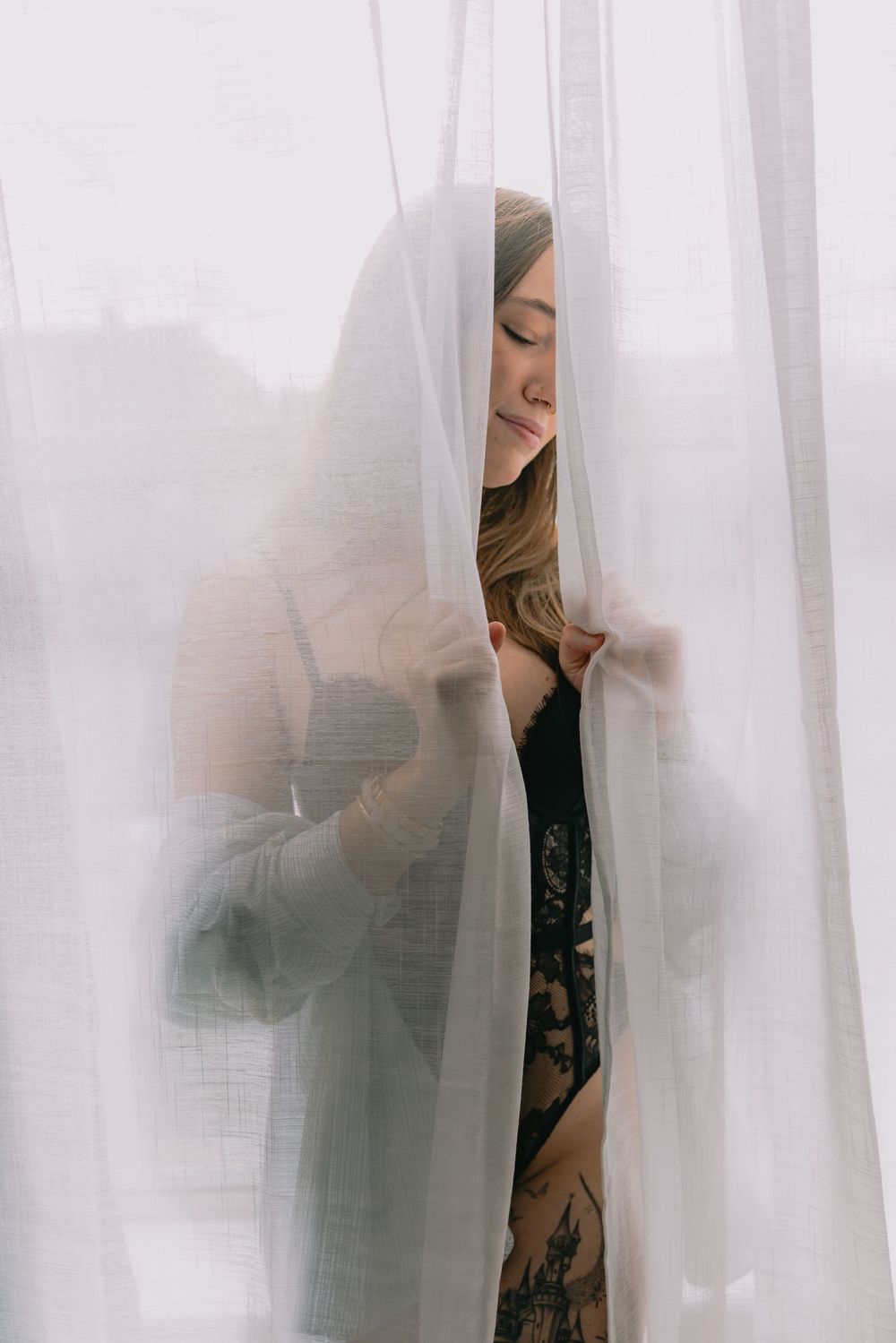 A dreamy portrait through sheer white curtains showing a figure in a dark outfit with a gentle expression.