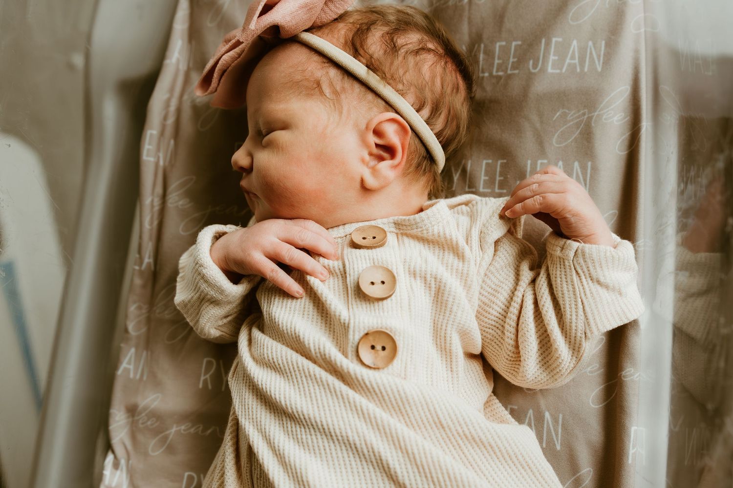 Sleeping newborn baby in ribbed onesie with pink bow headband on personalized blanket.