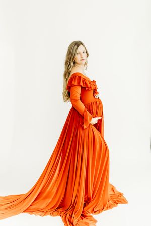 Woman in flowing bright orange maxi dress twirls gracefully against white backdrop in maternity photoshoot.