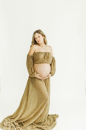 A pregnant woman in a flowing golden polka dot dress poses gracefully against a white background in a maternity photoshoot.