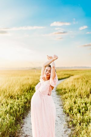 A person in a flowing pink dress twirls joyfully on a dirt path through a sunlit summer wheat field.